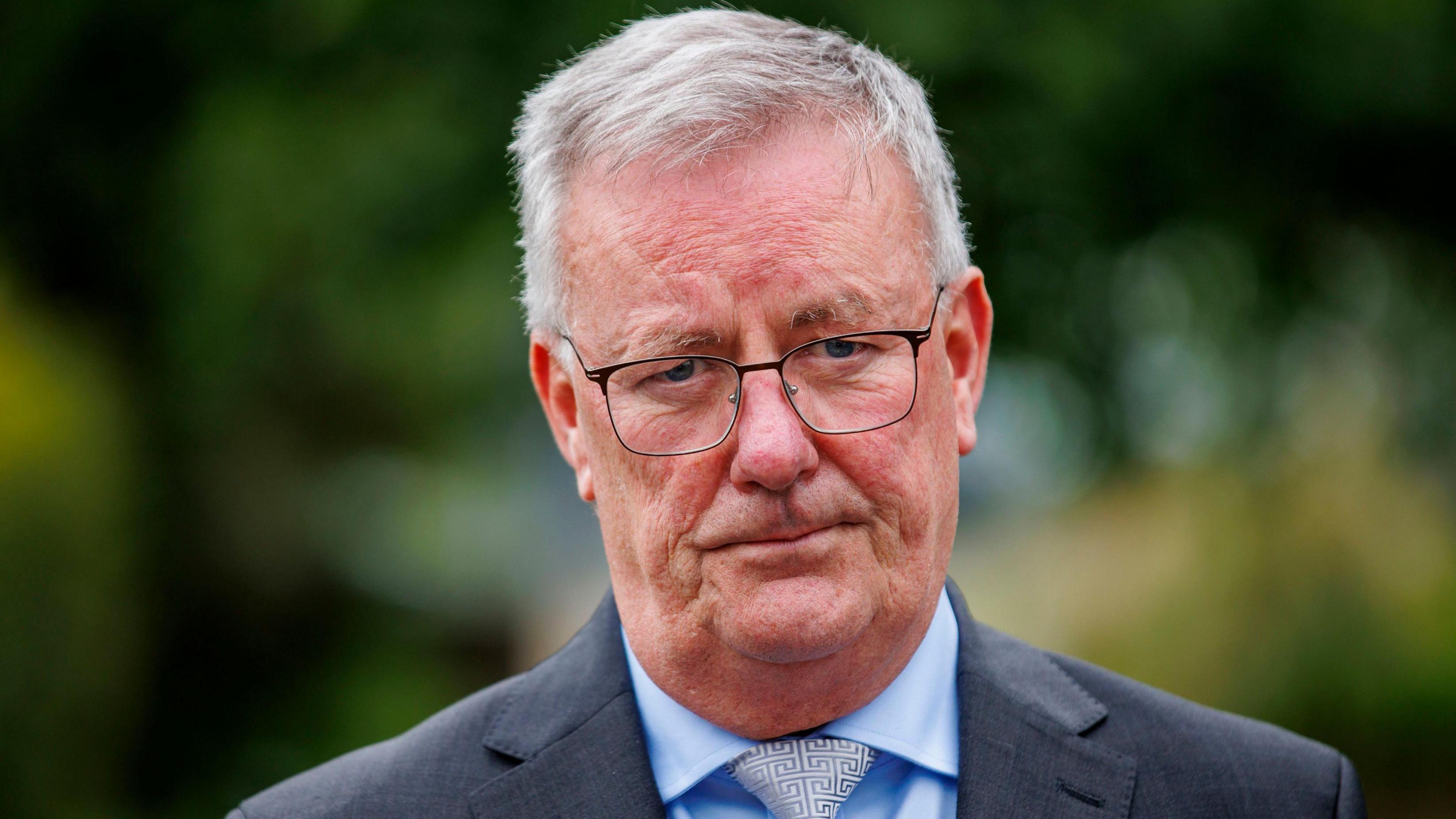 Mike Nesbitt is looking into the camera. He's wearing glasses and a grey suit with a blue shirt and a tie with a maze pattern. He's outside. The bushes in the background are out of focus.
