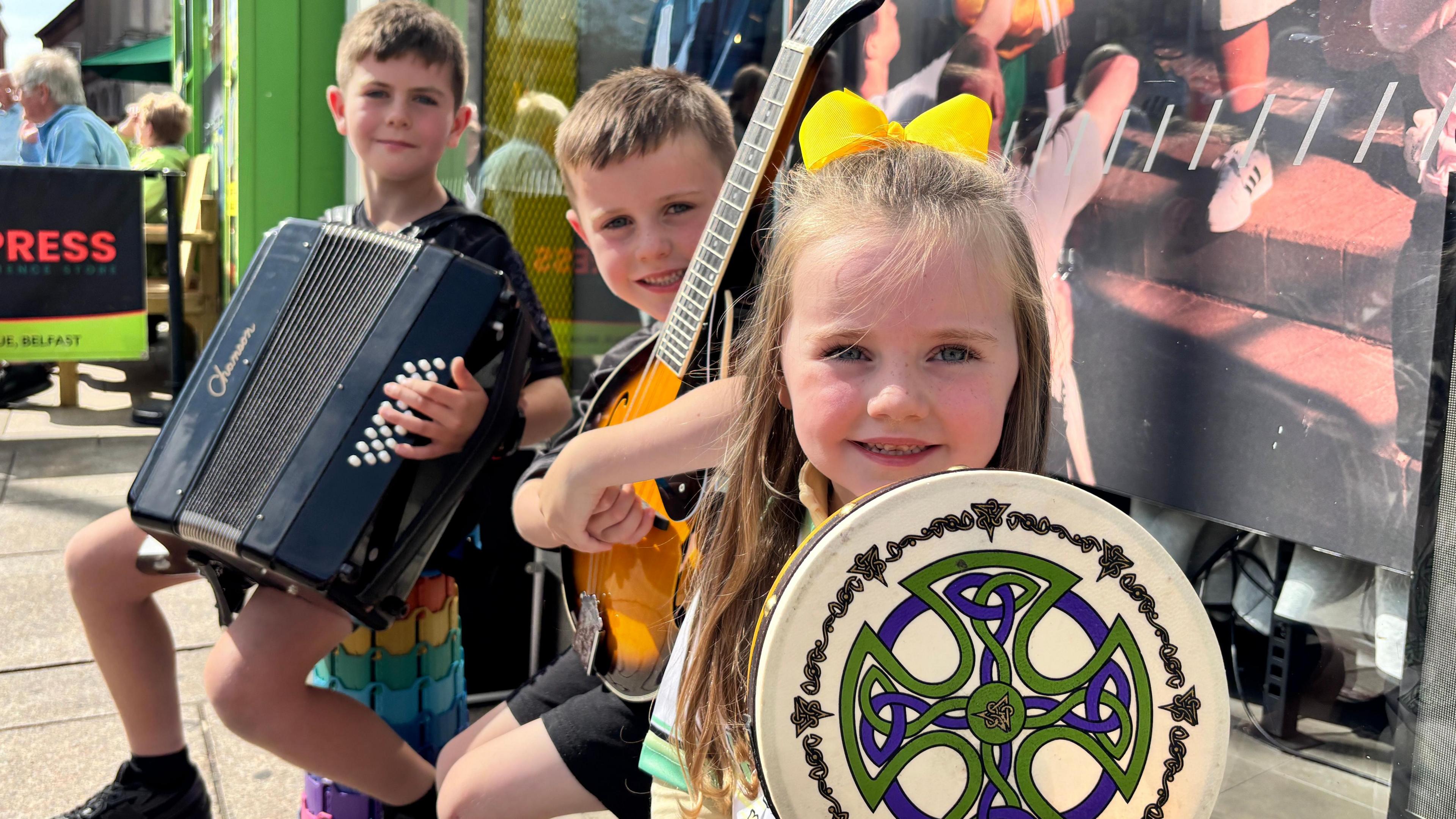 Two boys and a girl are holding instruments and smiling. One has an accordion, one has a guitar and one has a drum. There is a Celtic design on the drum. 