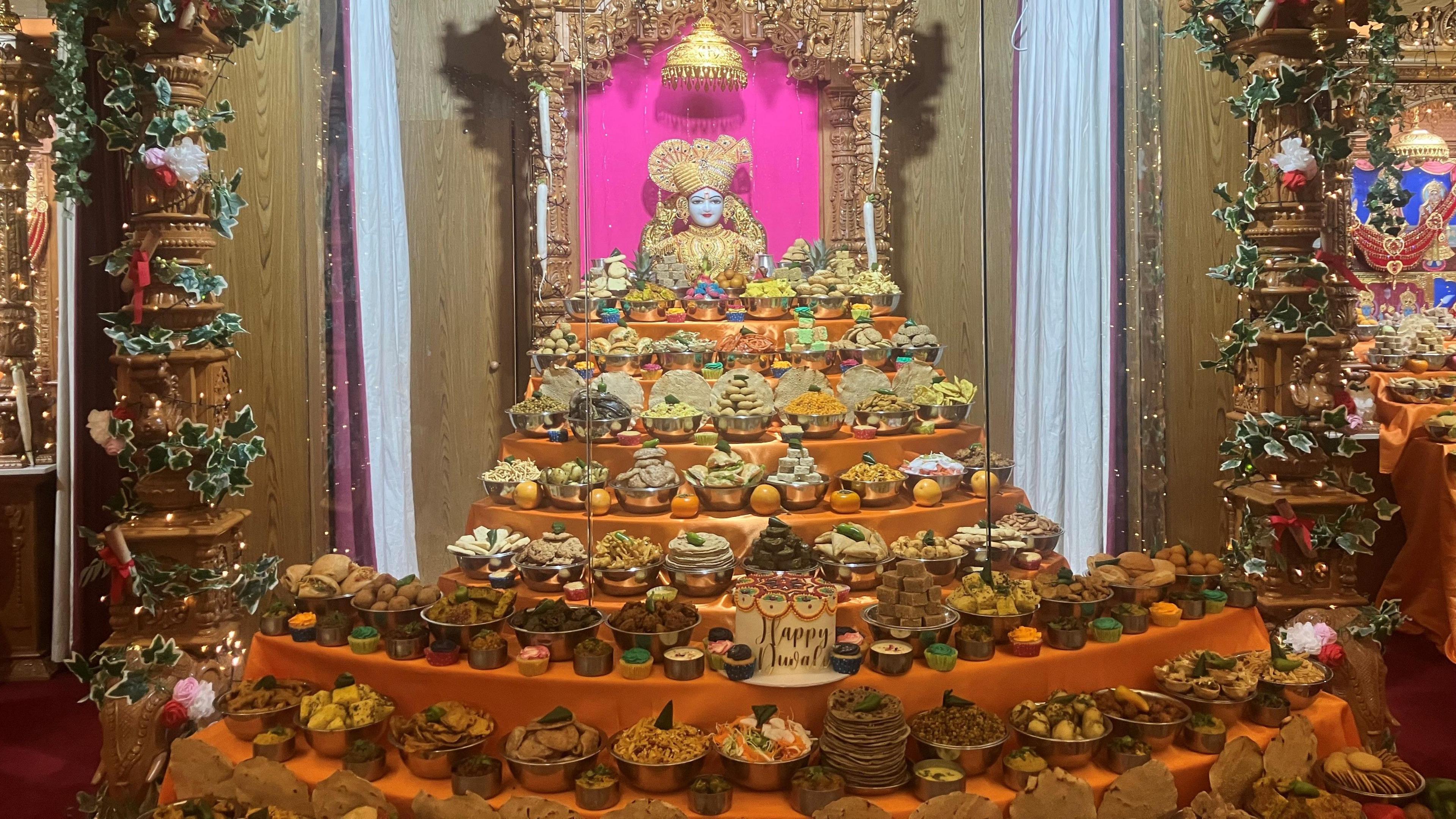 Bowls full of cakes and bakes goods are placed along a tiered orange arrangement. A statue of a Hindu deity is place in the centre of the stand.