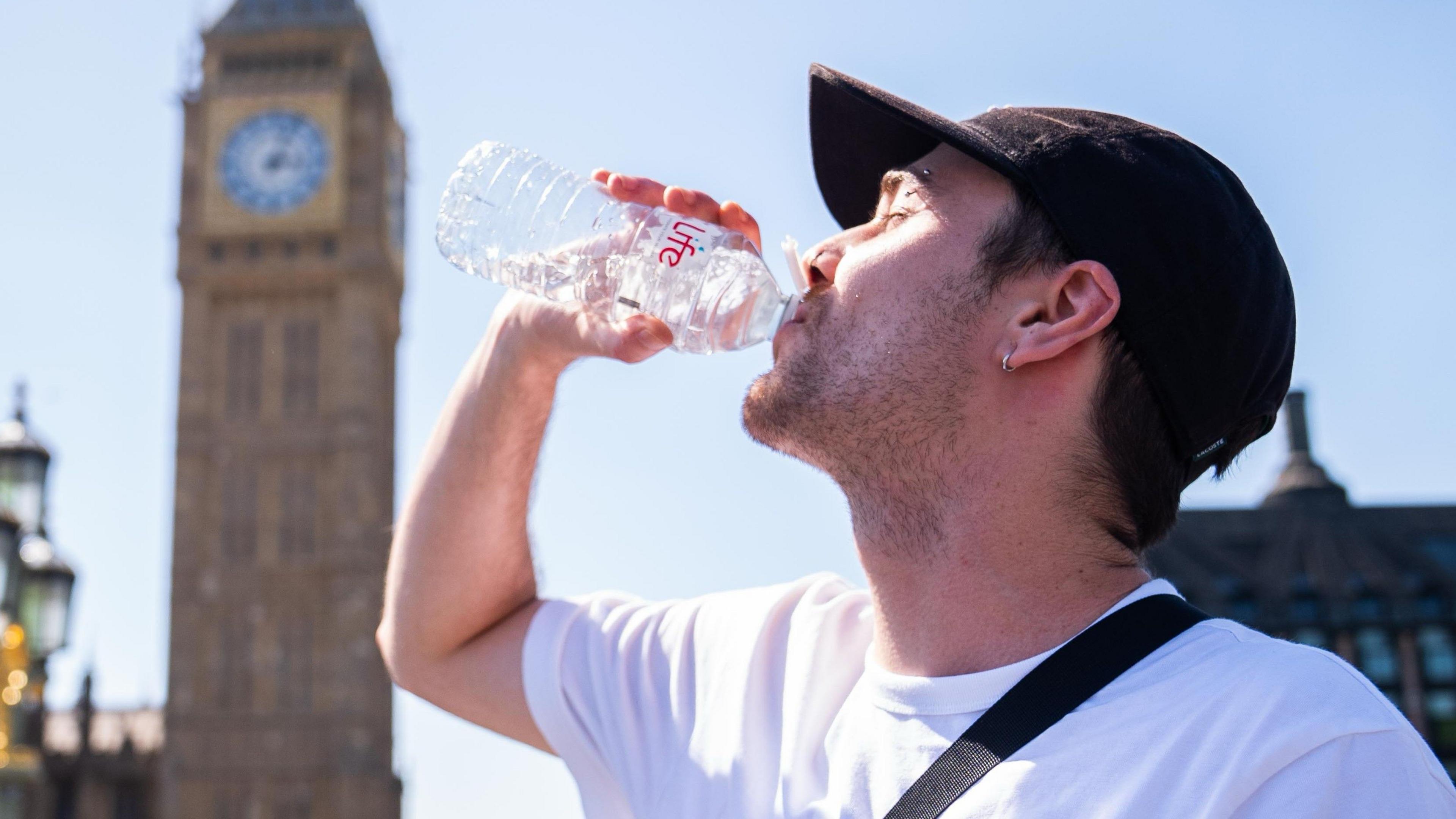 A man drinks from a water bottle with a sunny sky overhead and a blurred view of the Westminster skyline in the background