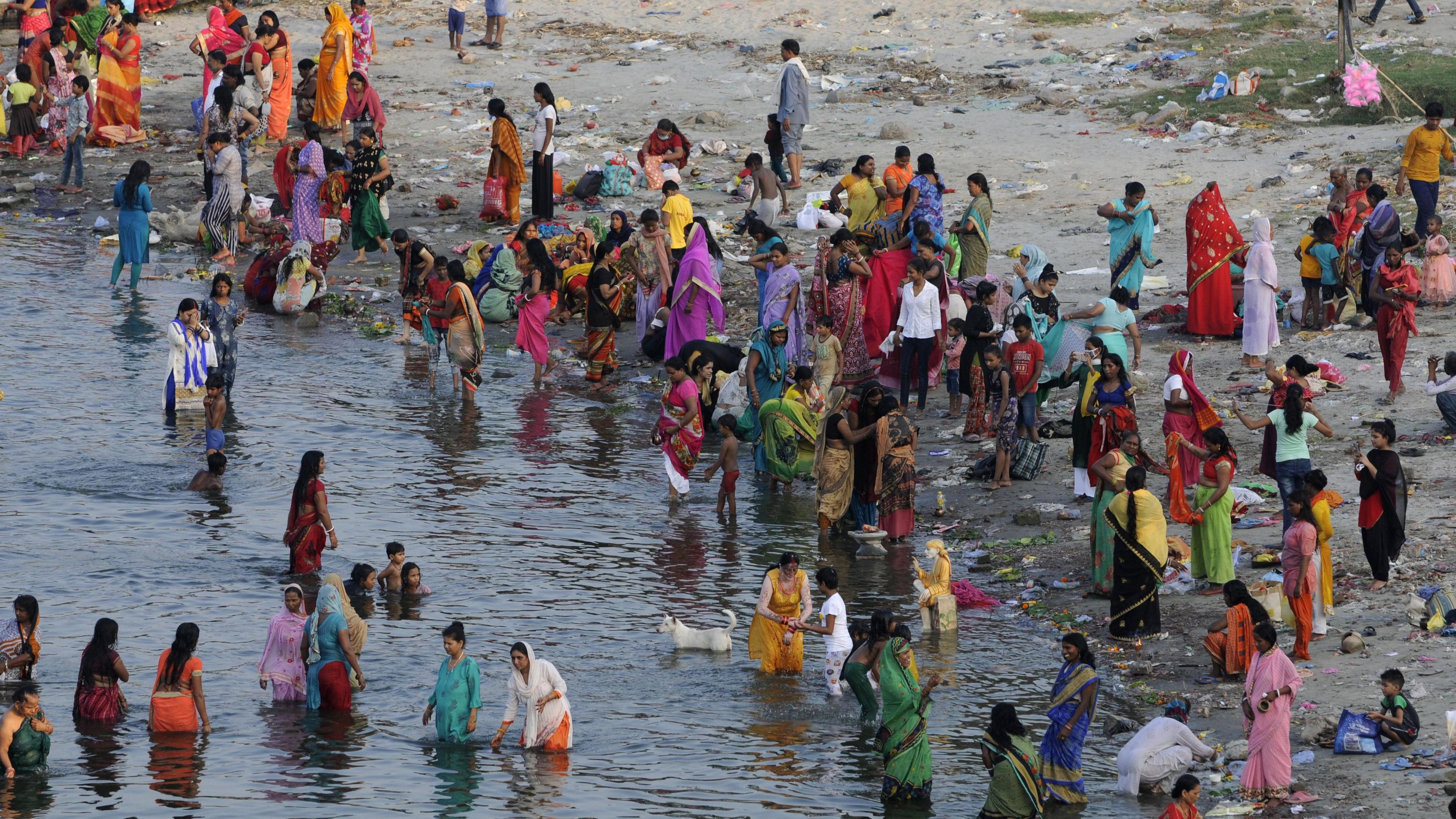 Women and children bathe in a river during the Jivitputrika festival in the Uttar Pradesh state in September 2020. The water is quite dark, in contrast to the many colours of the clothes that people are wearing. The sand along the bank is grey and there is a lot of rubbish strewn everywhere.