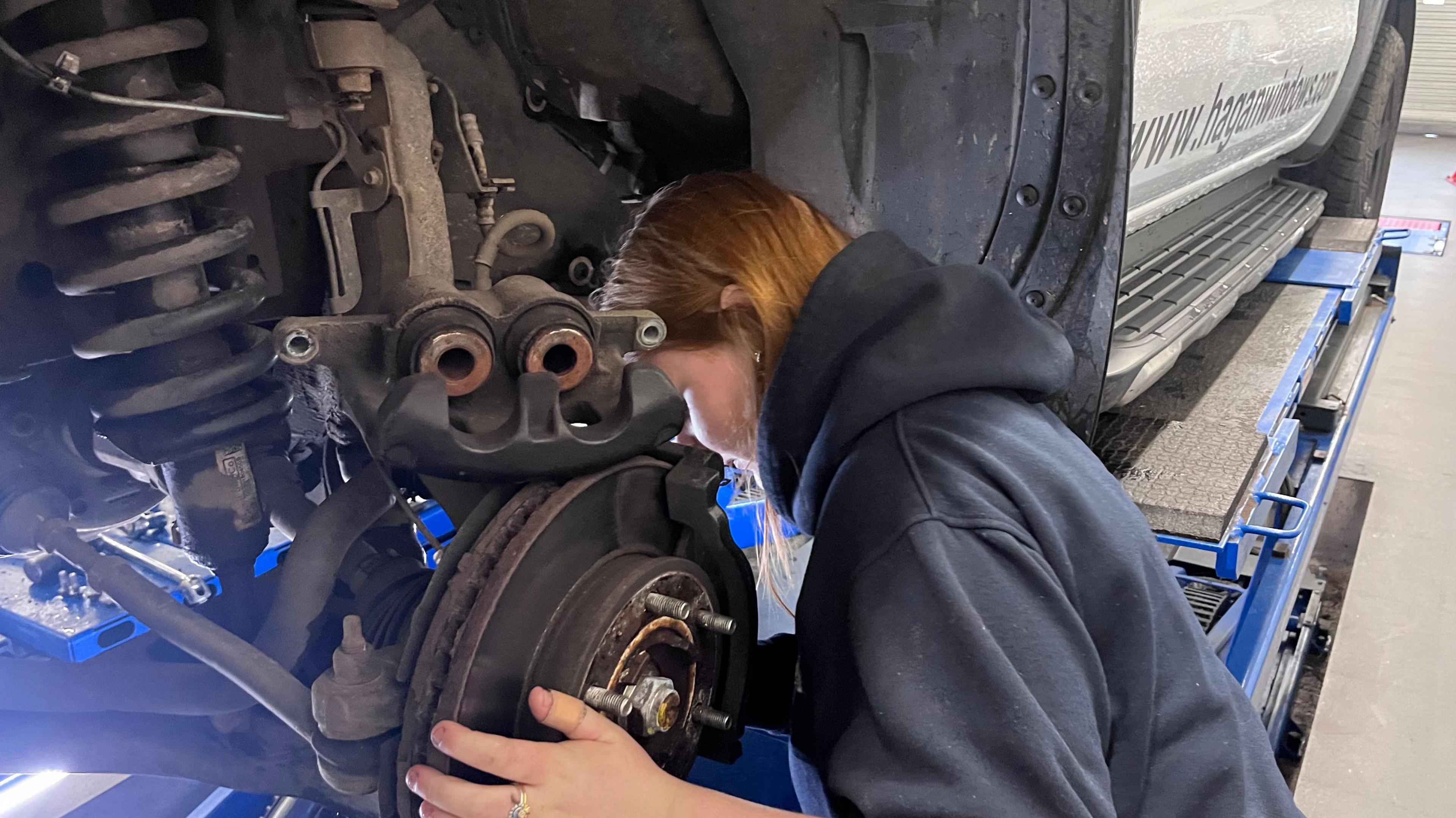 A close up of a female with ginger hair and a grey jumper who is replacing a car tyre.