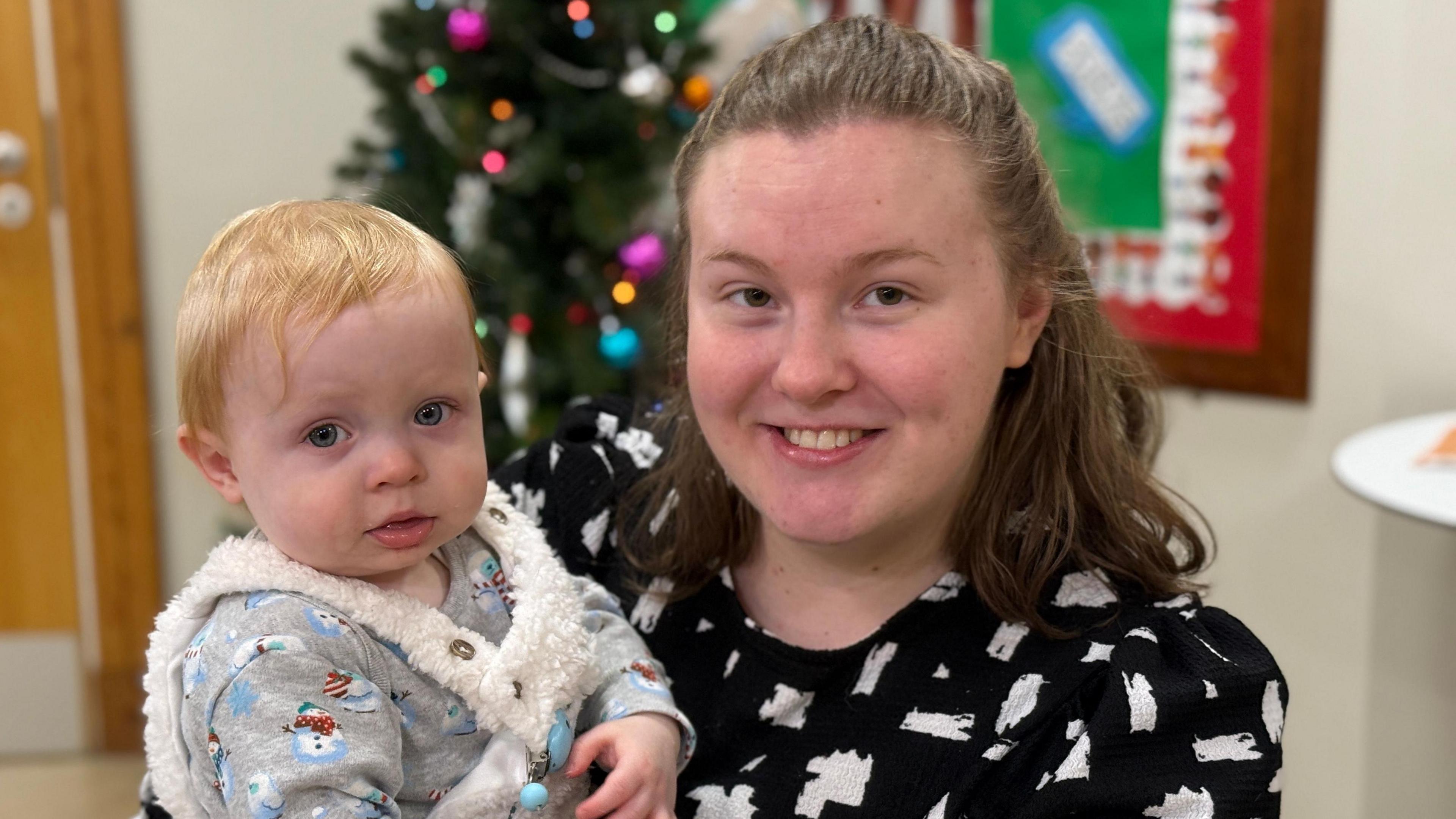 Robin smiling and holding a child in her arms. They are both looking at the camera. She is wearing a black-and-white top. There is a Christmas tree in the background.