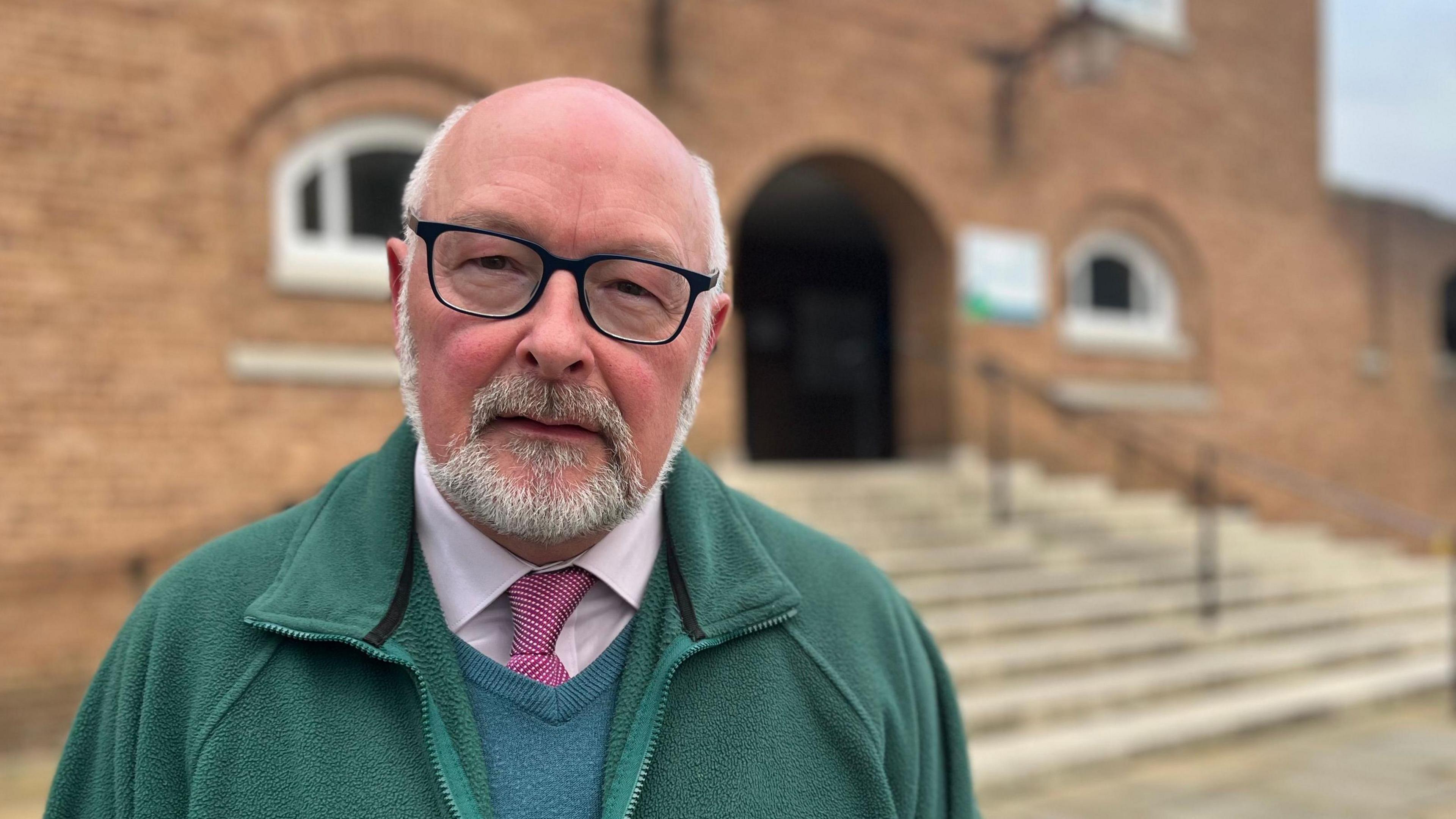 Alan Connett, Liberal Democrat councillor on Devon County Council, wearing glasses and a pink spotted tie, teal sweater and green fleece, standing in front of County Hall