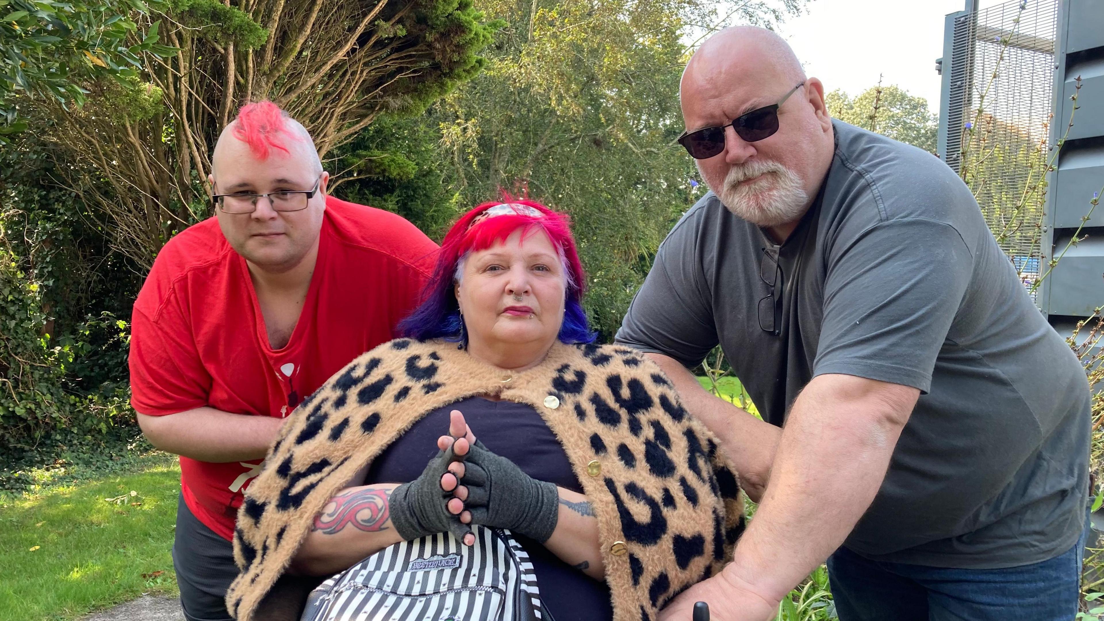 Jake Hughes, Karran Minchinton and Sean McEvoy are in a garden looking at the camera. Mr Hughes is standing and has a pink mohican and glasses, Ms Minchinton is sitting and has red and blue hair and is wearing an animal print cardigan and Mr McEvoy is standing and has a goatee and is wearing sunglasses