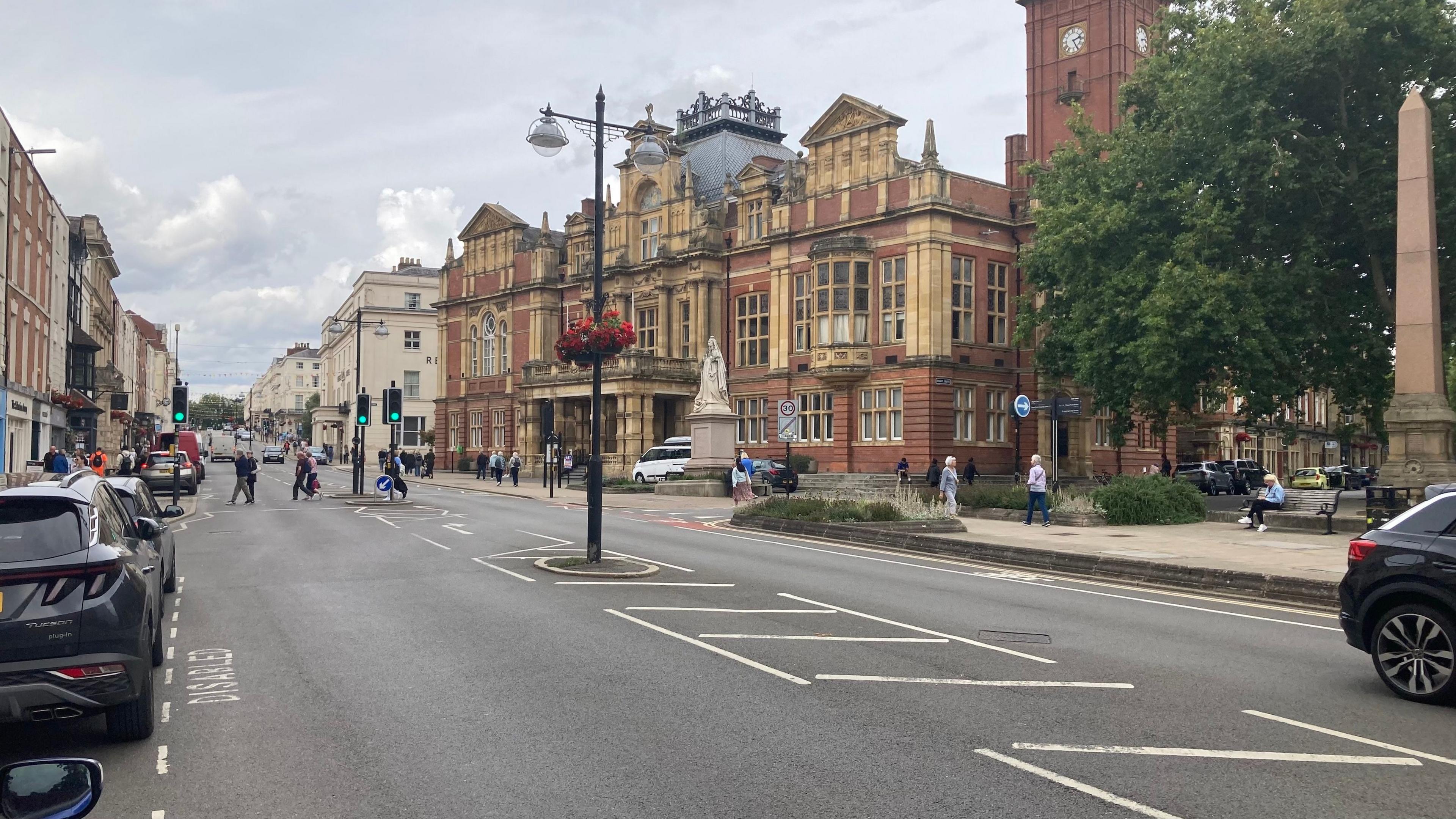 A high street with old buildings on either side, some cars parked on the left, showing and people crossing the road further down. 