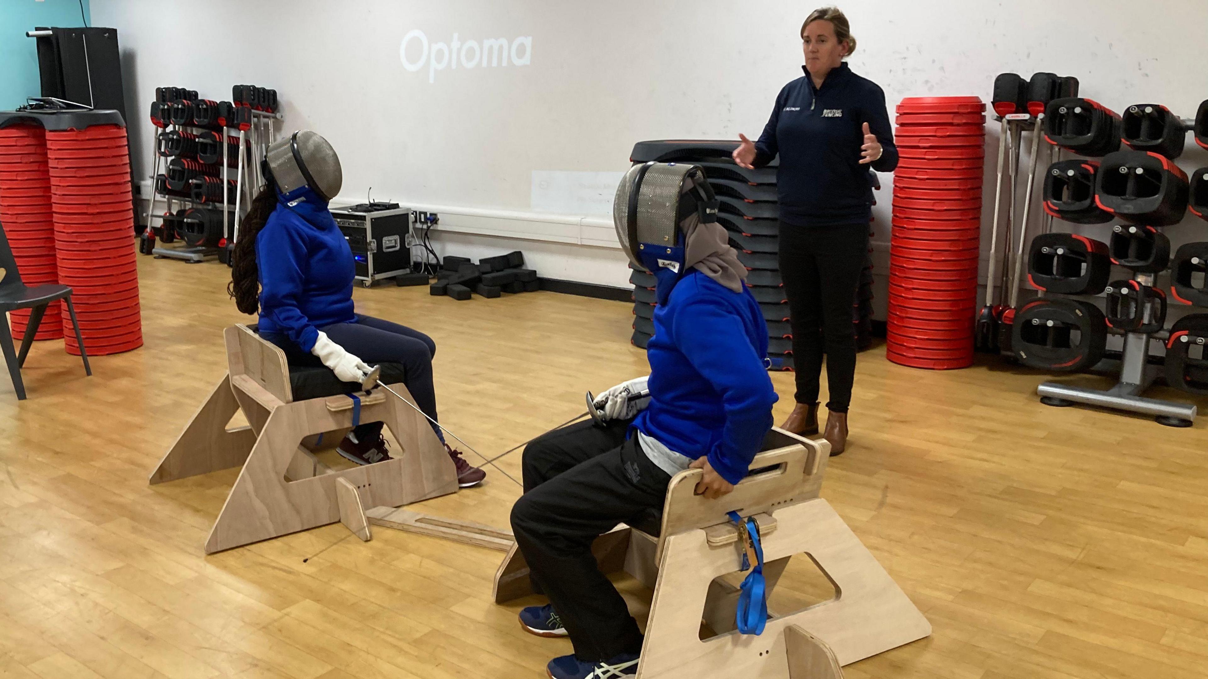 Two group members try out the sword seats. They are both wearing masks and gloves and are holding swords and fencing and listening to an instructor.