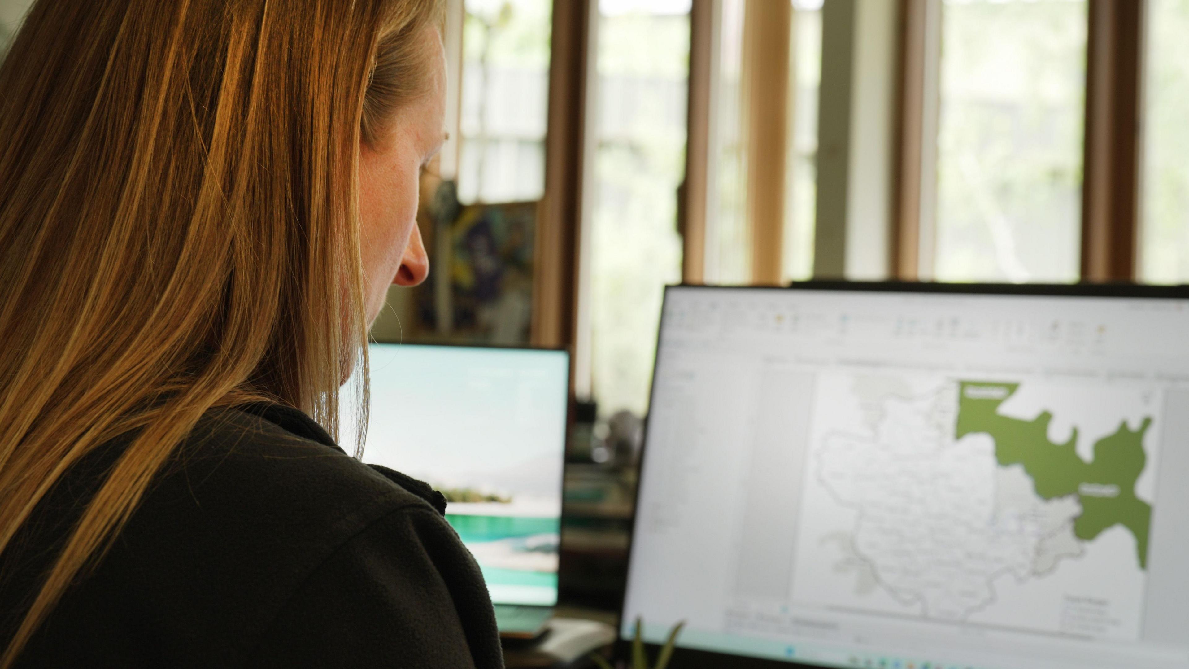 A woman with long blonde hair looks at a computer screen with a map on it.