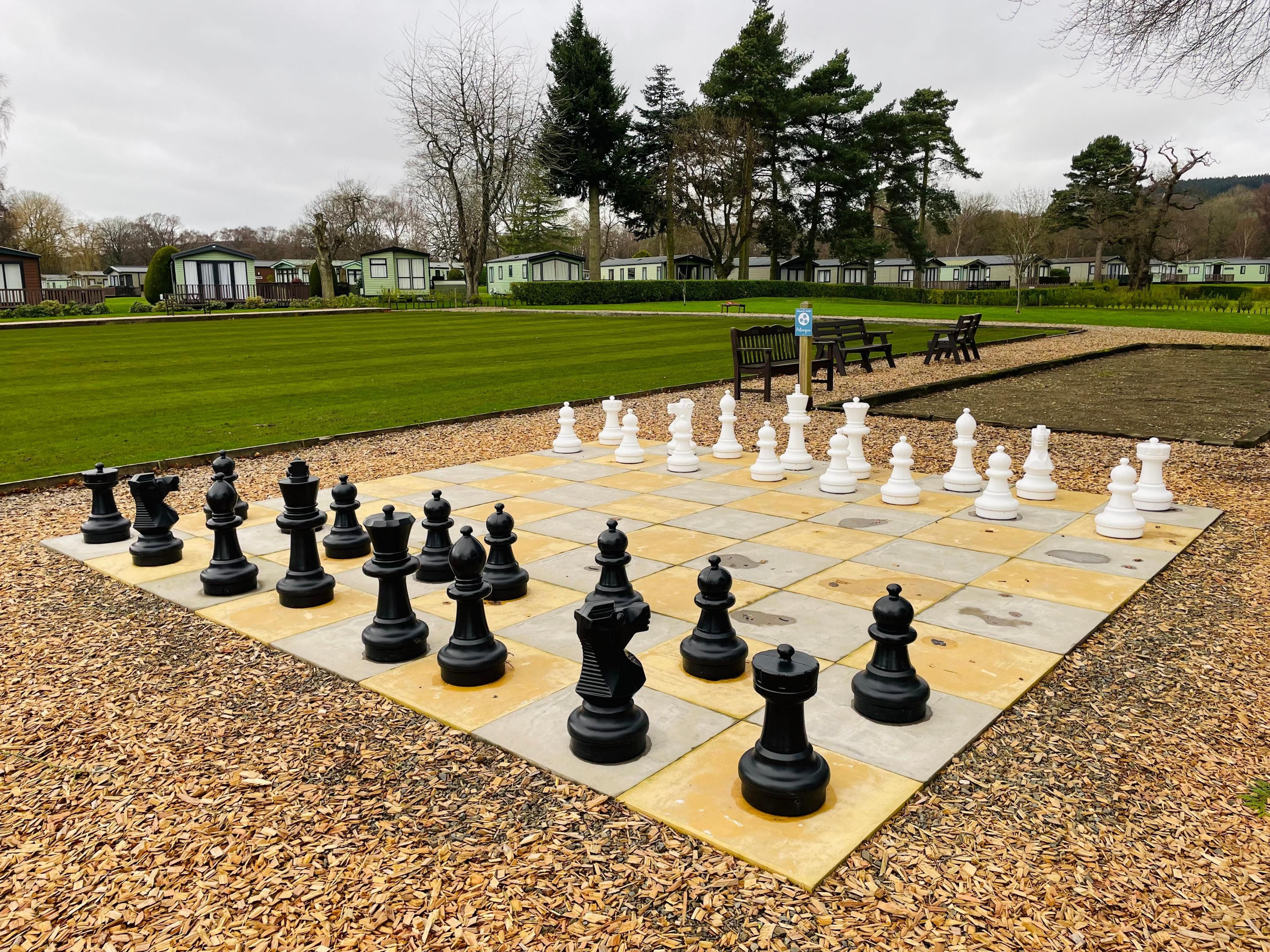 A chess board laid out in golden and grey paving slabs on a surround of wood chippings, with black and white chess pieces laid out for the start of a game. Static caravans are visible in the distance.