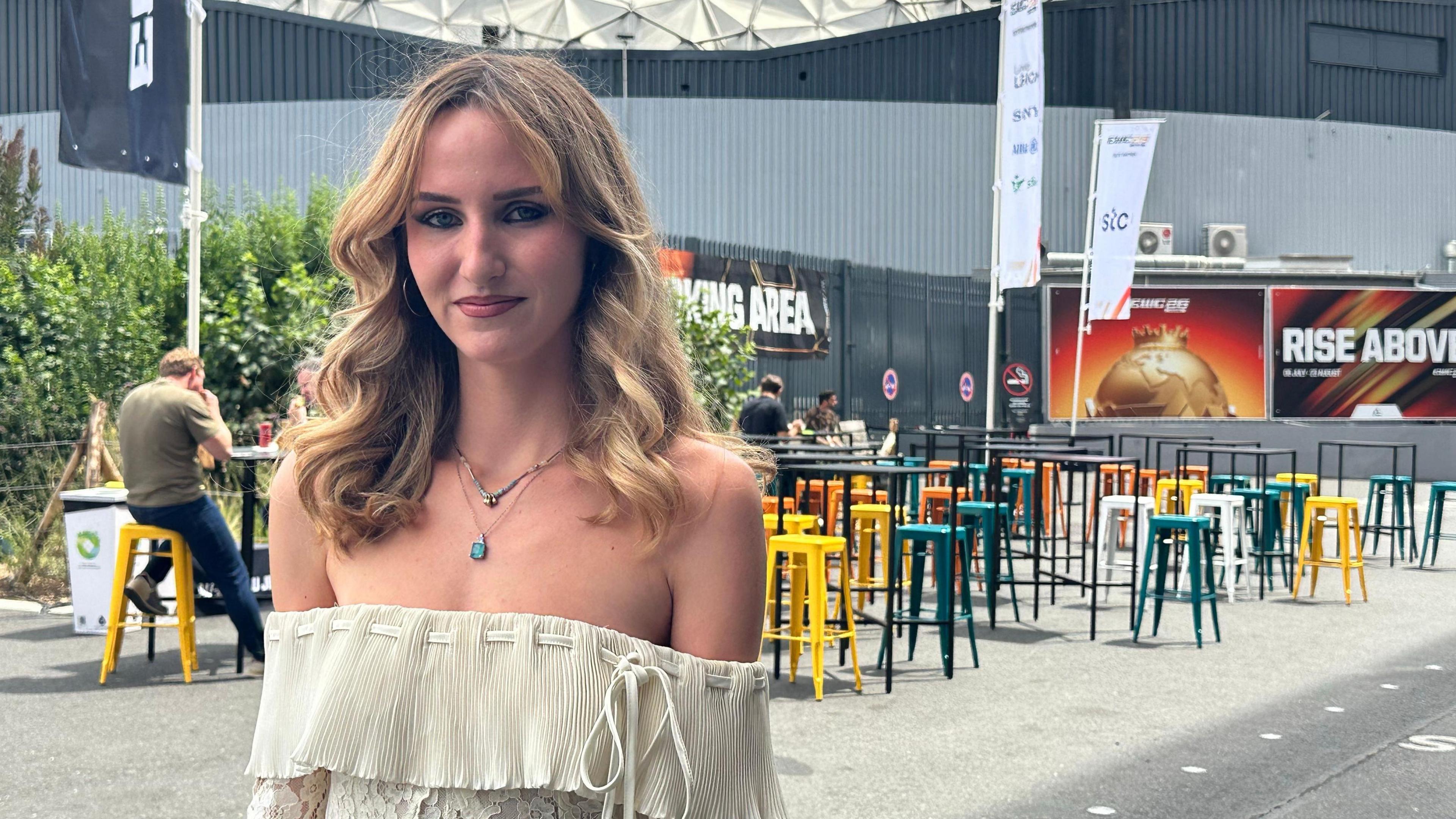Jenny has curly blonde hair which sits on her shoulders. She wears a cream floating top with a turquoise necklace