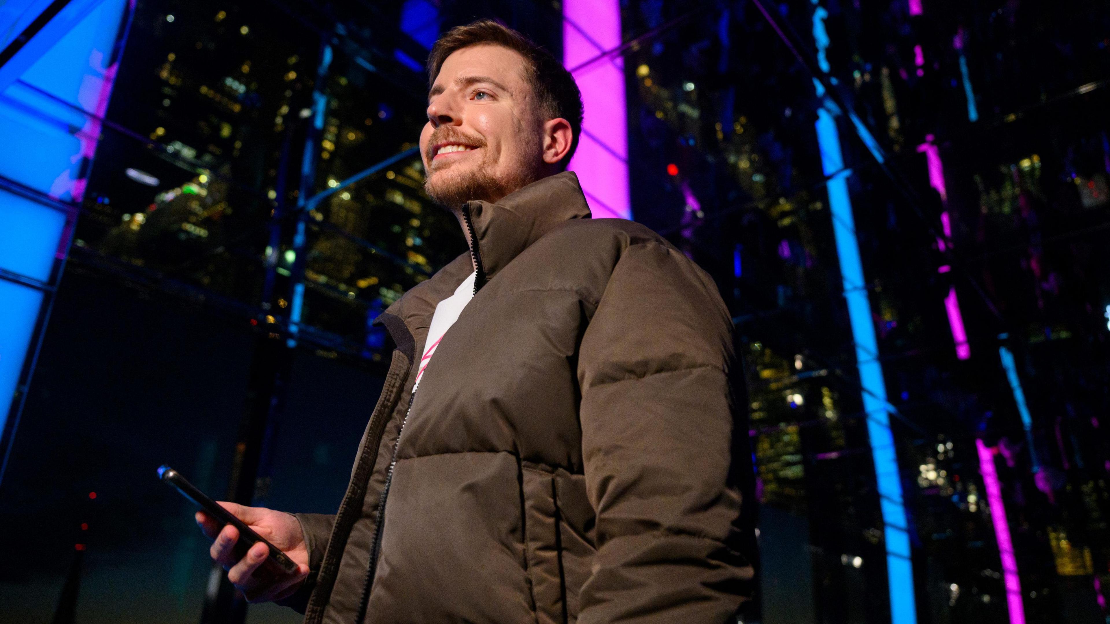 MrBeast pictured outside in New York on a  December evening in front of a glass building illuminated in blue and pink to promote Beast Games. He wears a brown puffer jacket and holds a smartphone in his hand. 