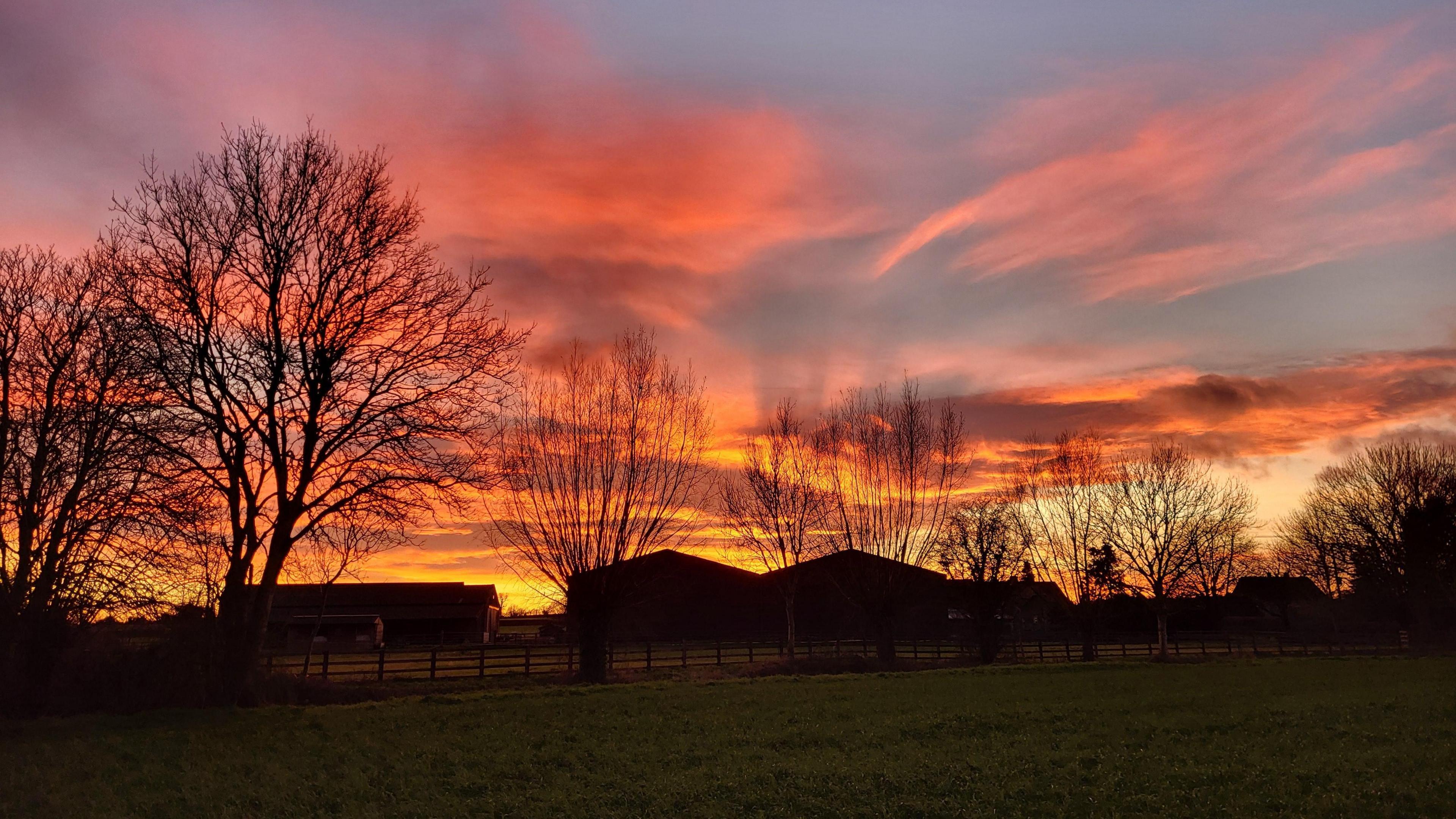 The sky is a mixture of yellow, orange and pink. Beneath it sits a green field and wooden fence with several large farm buildings behind the fence.