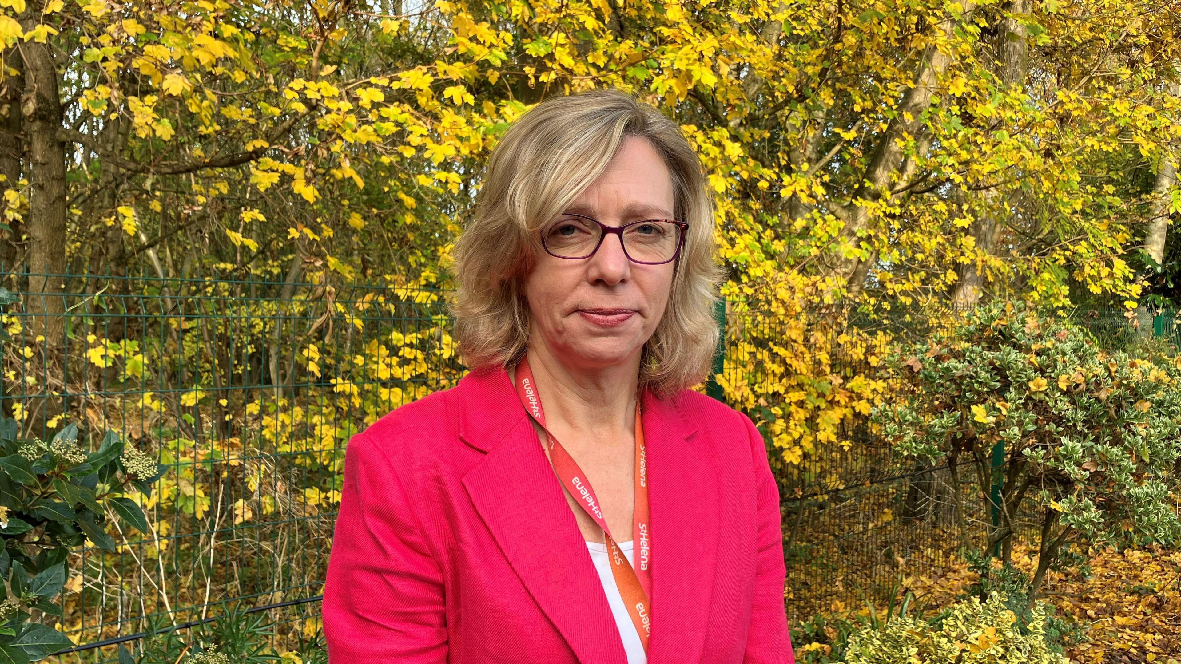 A head and shoulders picture of Dr Karen Chumbley standing in front of some trees. She is wearing a pink blazer.