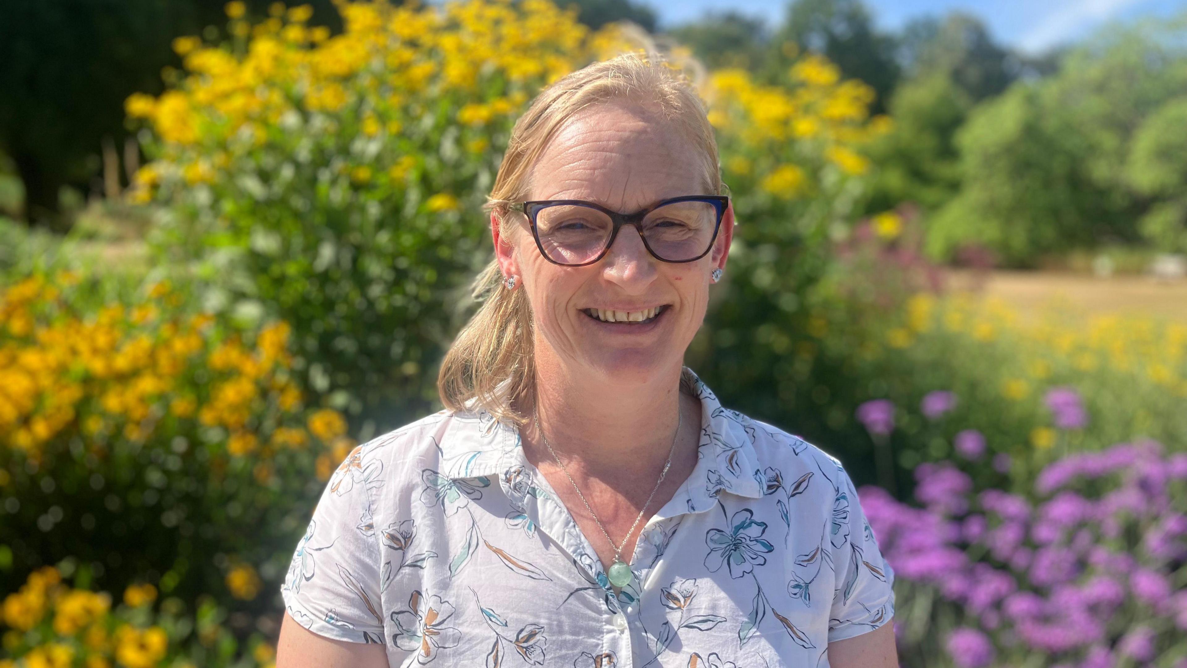 A woman wears a white shirt and glasses. She has blonde hair and stands in front of a herbacious border which is blurred behind.