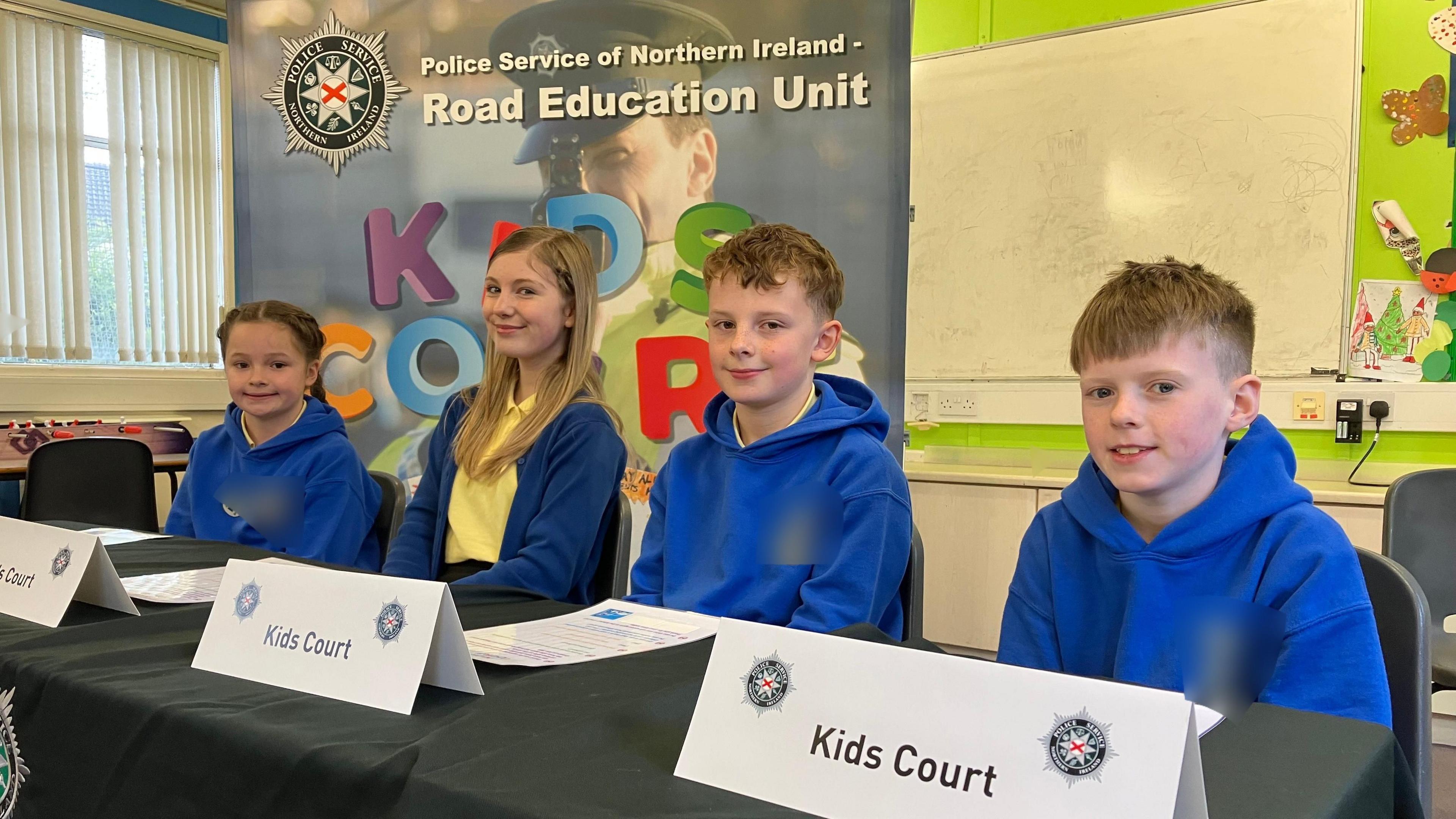 A number of schoolchildren, two girls and two boys sitting at a table in a classroom. There are several signs on the table with PSNI symbols which say 'Kids Court'. The uniform is royal blue and yellow.