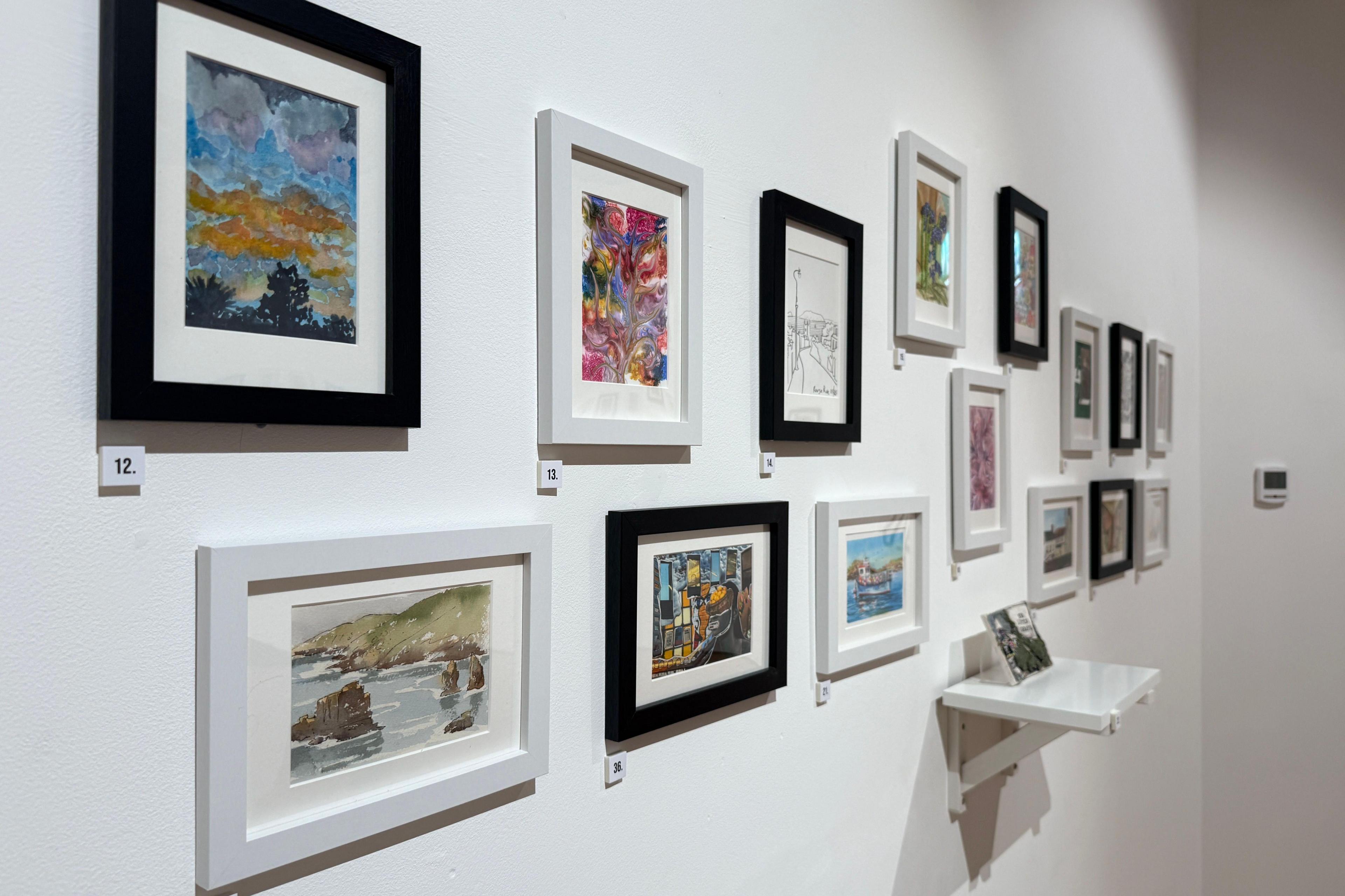 A picture at an angle showing 13 frames with art in them . There is a small shelf in the middle of the display with a ceramic piece.
