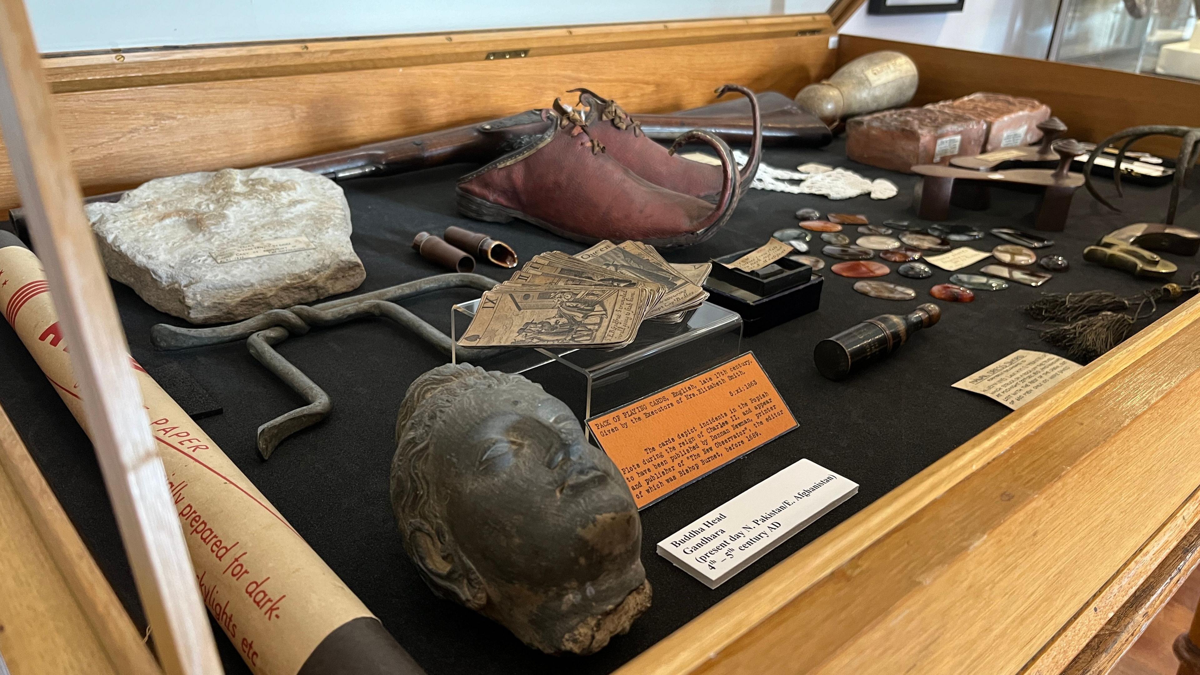 Items are on display in a cabinet, including a sculpted head, a pair of brown shoes with curled spikes on the toes, a collection of colourful stones and some playing cards.