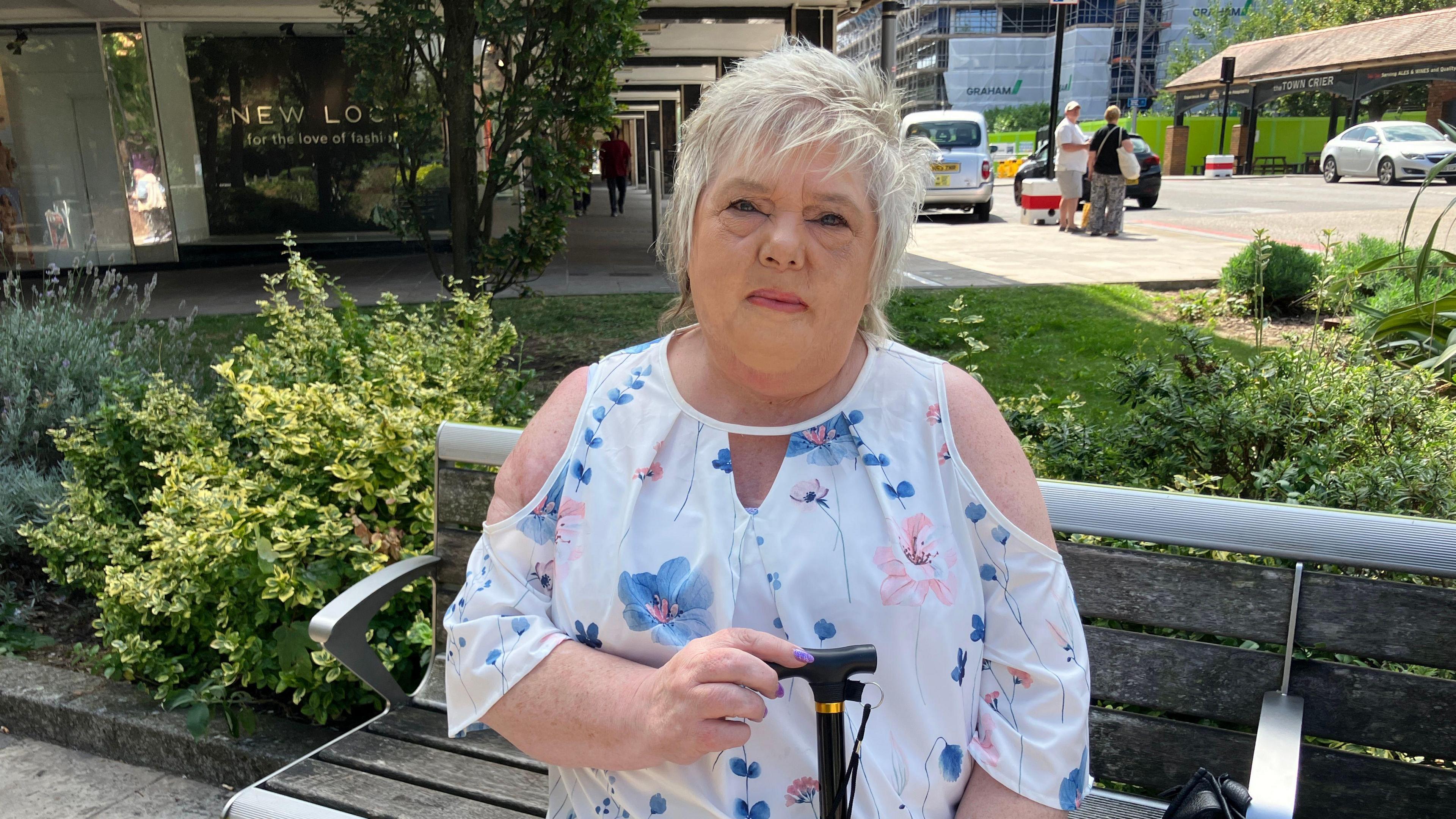 Pearl Miller wearing a white floral shirt sat on a wooden bench holding a black cane