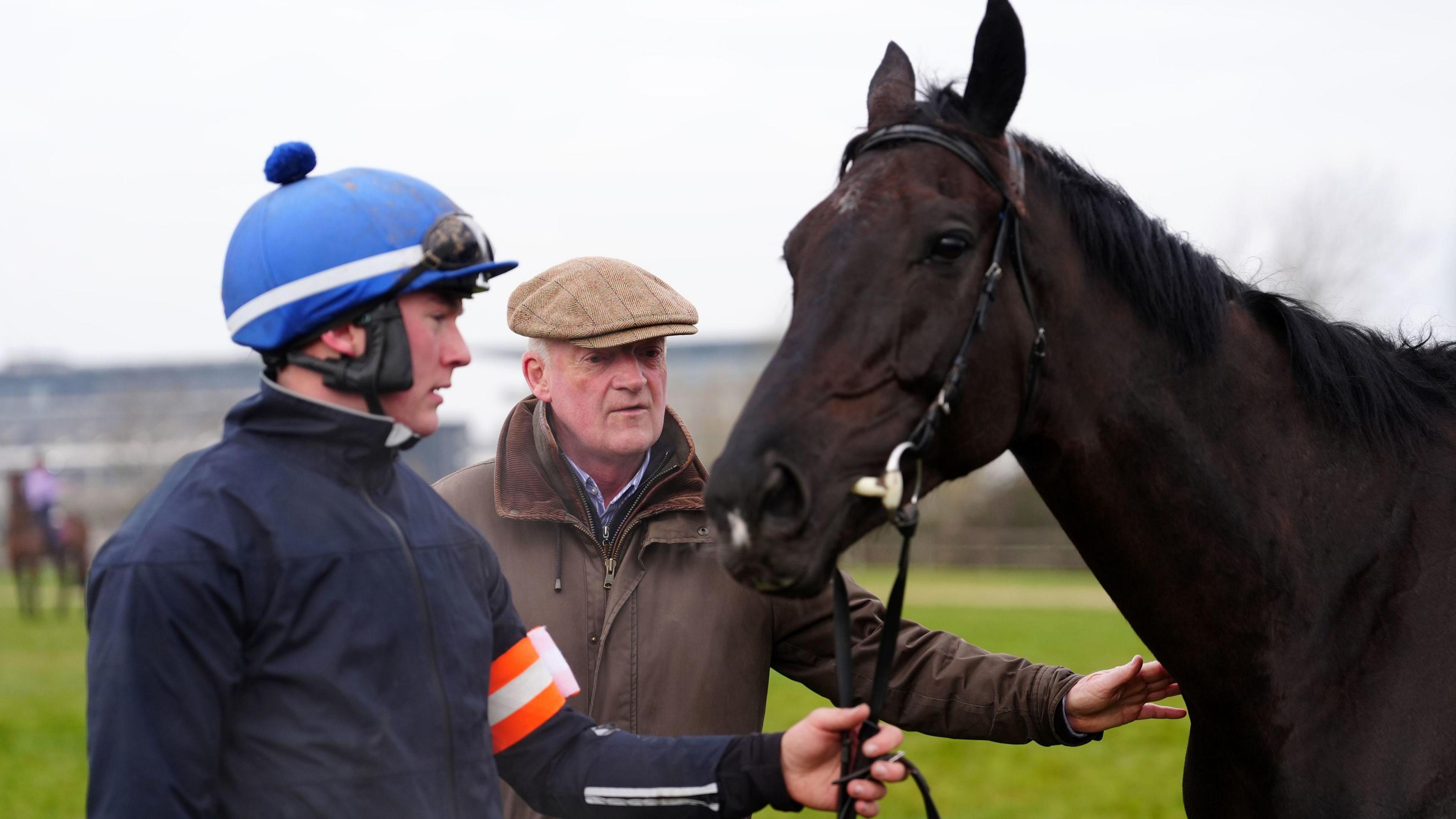 A jockey wearing a blue helmet and dark blue clothing holds the reins of Galopin Des Champs, a dark brown horse, as they stand on a field. Galopin Des Champs' trainer, Willie Mullins, stands behind them.