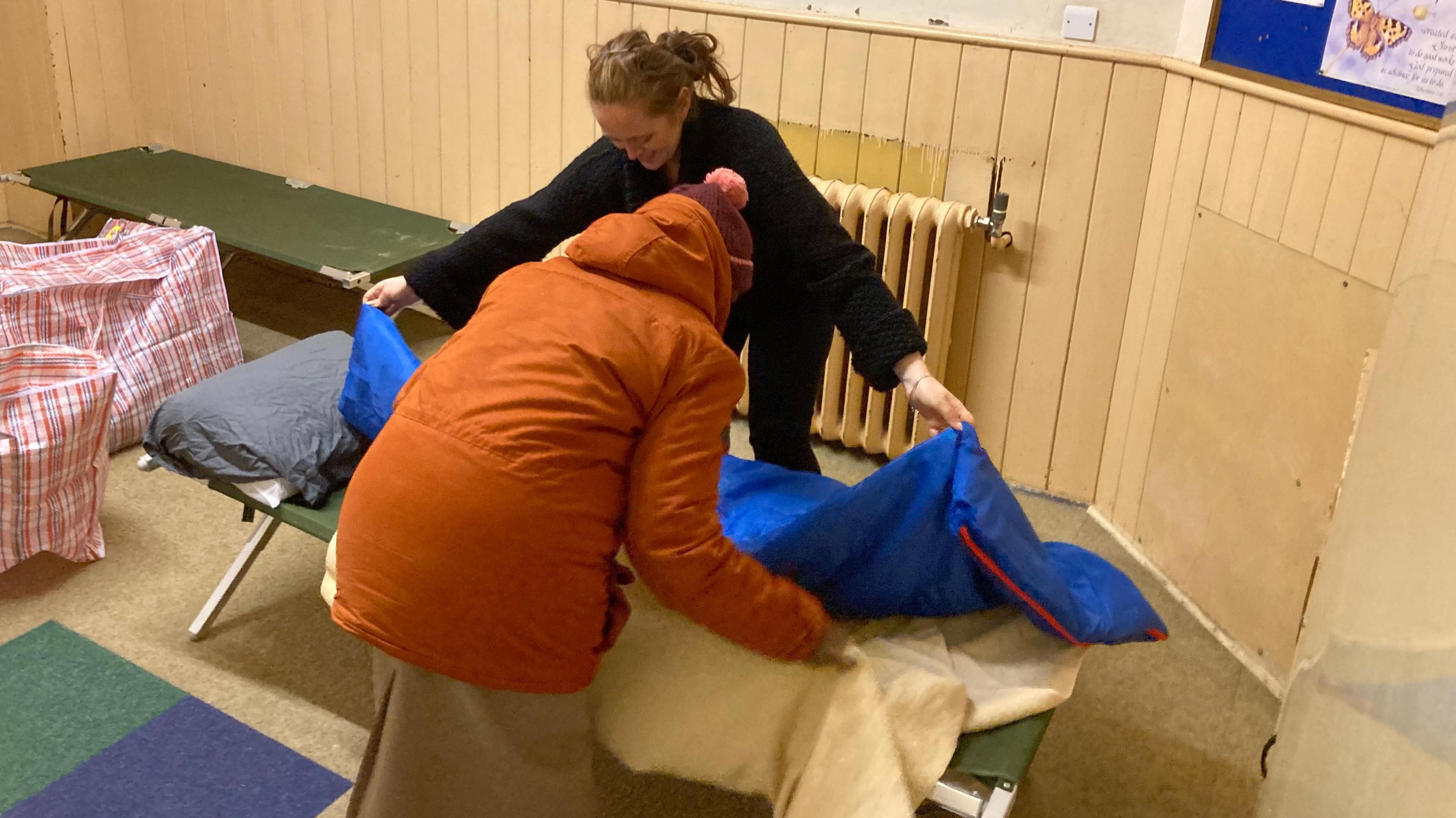 One woman dressed in black making up a bed at a church hall alongside another person dressed in an orange coat and hat. 