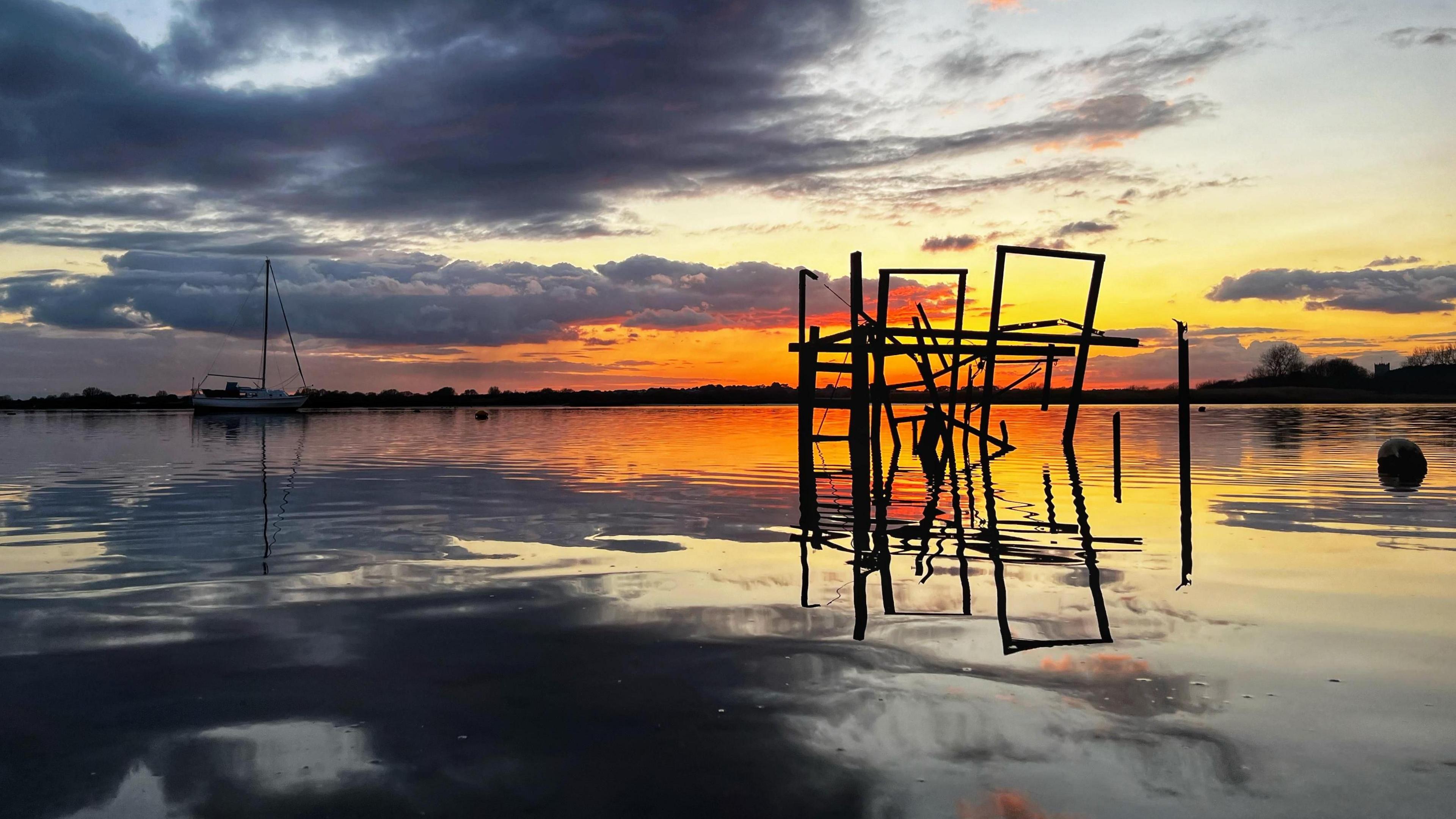 A vivid orange sunset over the harbour is reflected in the water. The sky has dark clouds above the orange/yellow glow. In the water you can see a sailing boat and a collapsed structure. 