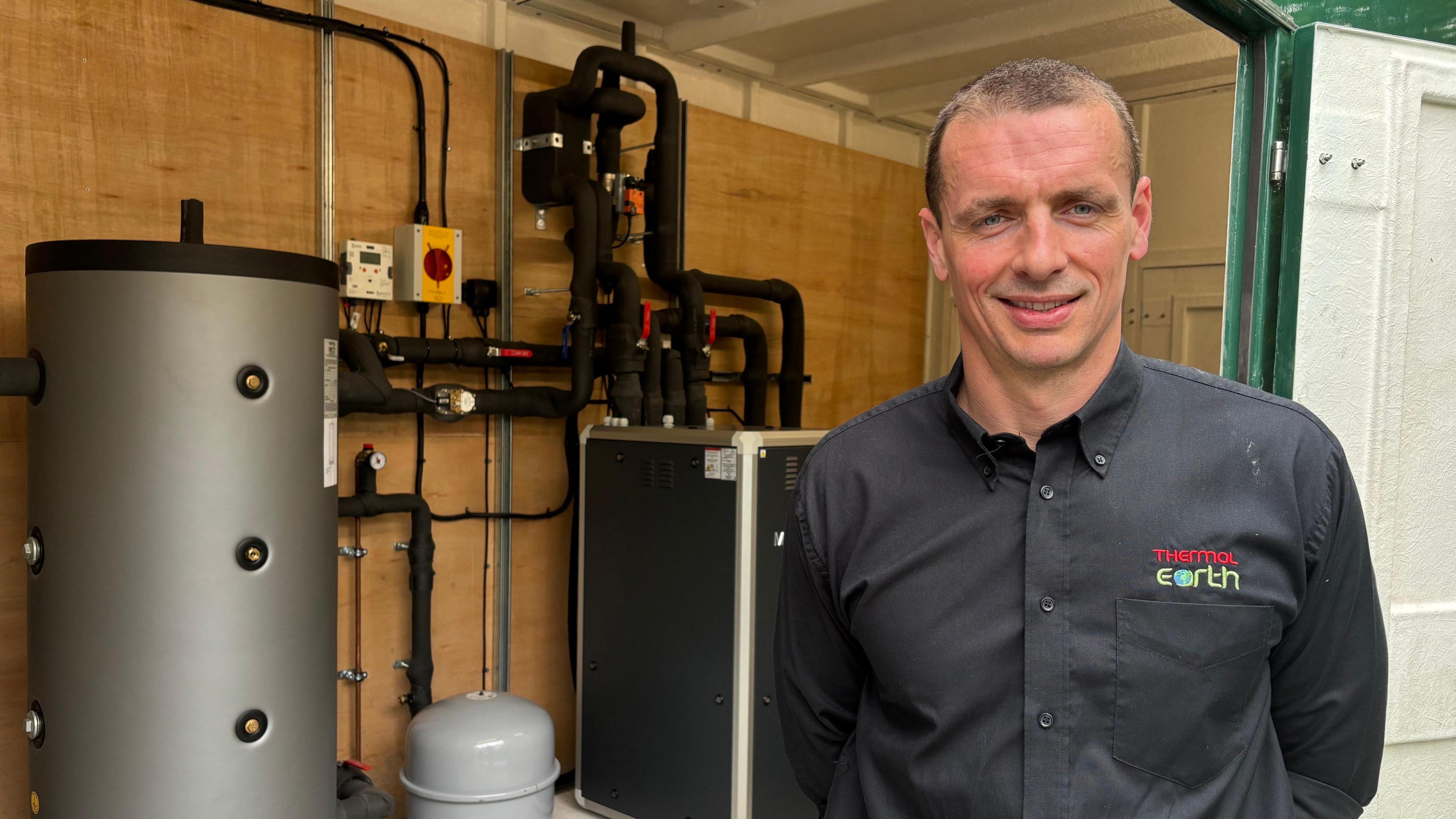 Nick Salini with short hair and wearing a black shirt, standing right of frame. Behind him is a heating system with black pipework, a compressor and boiler system.