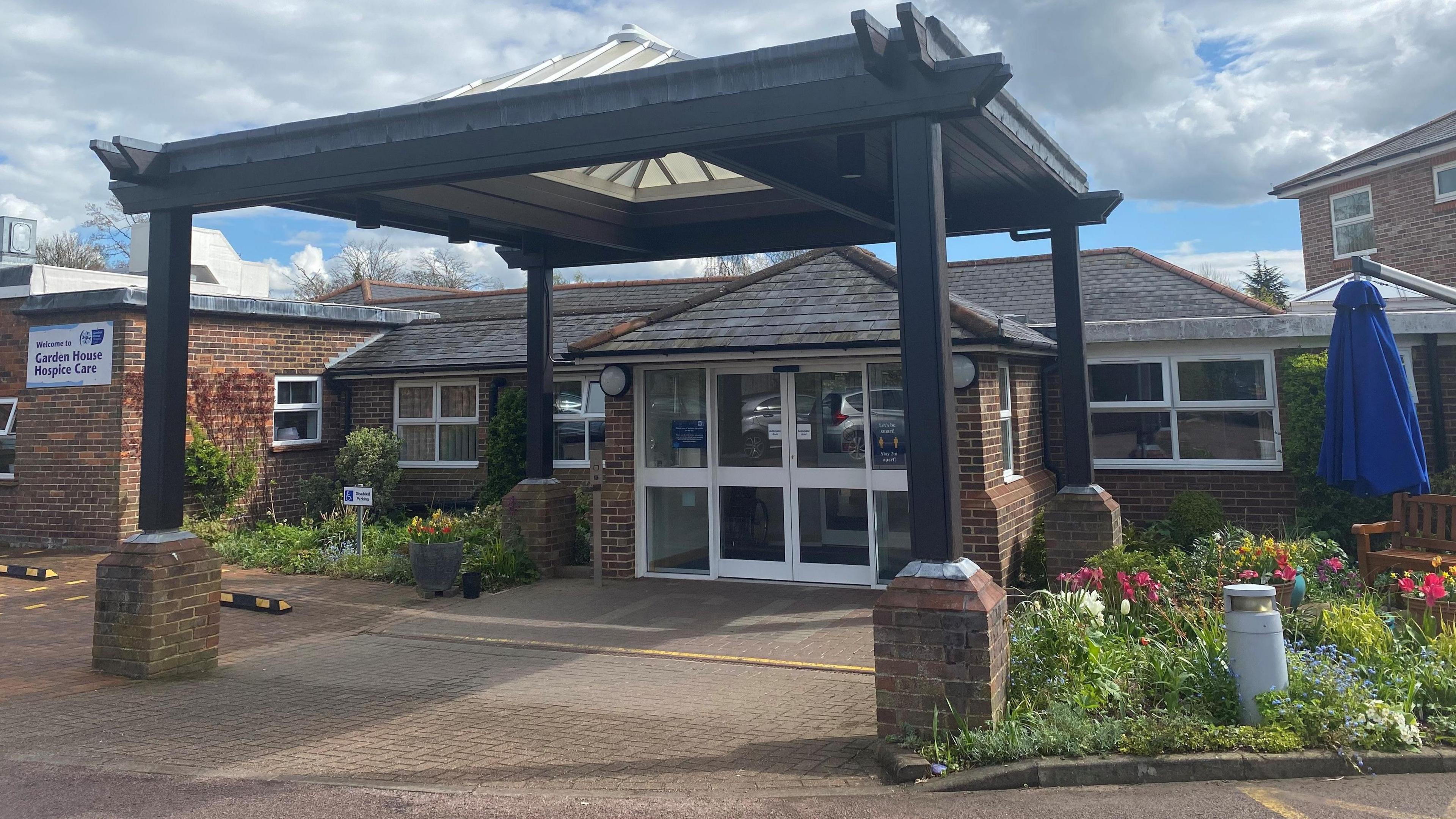 The outside of Garden House Hospice, a single-storey brick building with sliding doors at the porch entrance. There is a pergola in front of the porch with a pyramid translucent roof. To the right is a small garden with flowers, a wood bench and blue parasol. 