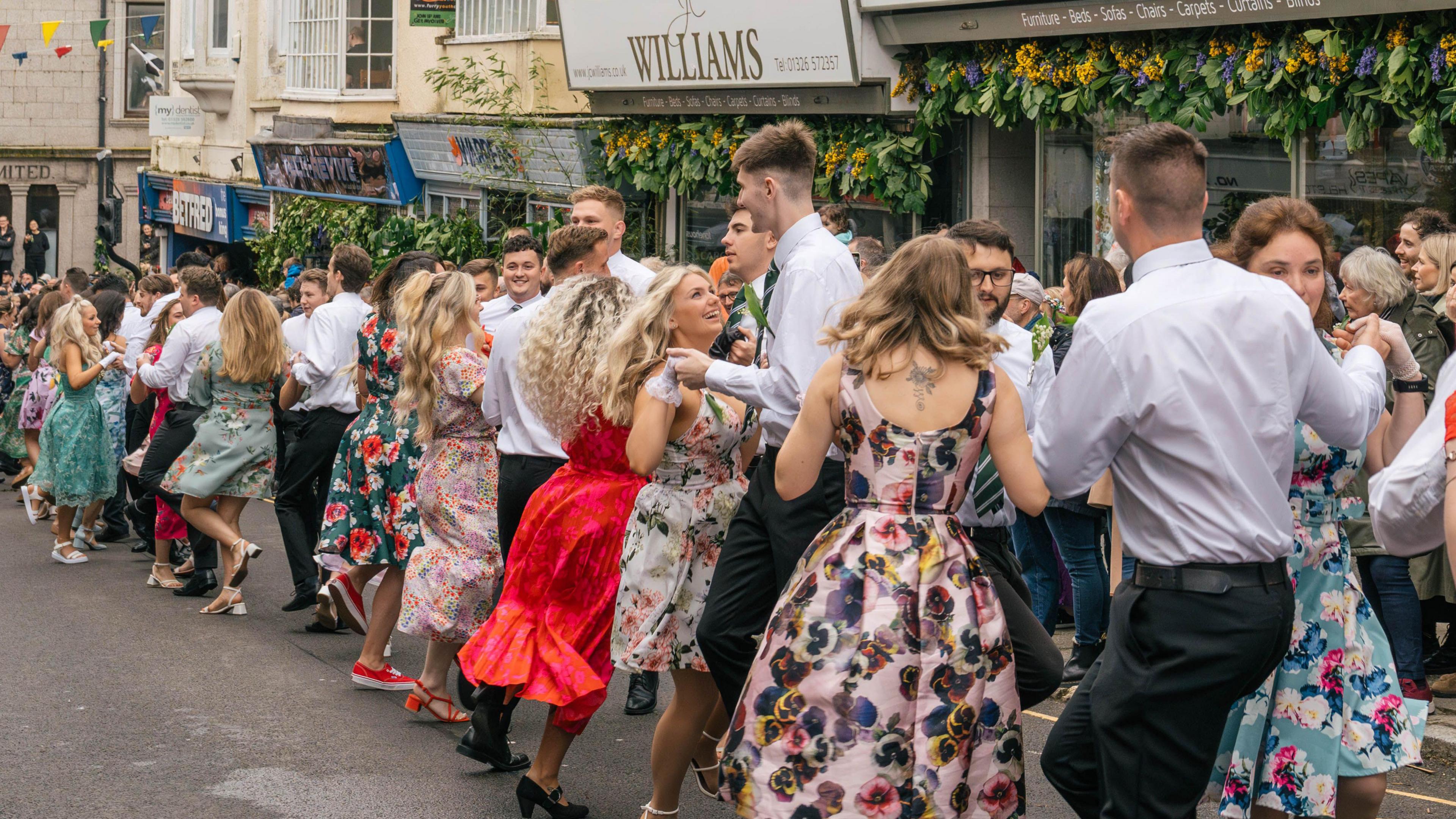 Young men and women are dancing in the street. The women's mid-length dresses are brightly coloured and many have floral designs. The women are wearing gloves. The men are wearing white shirts and black trousers with green and yellow striped ties. The shops lining the street are decorated with sunflowers, greenery and bluebells. There are spectators on the pavement.