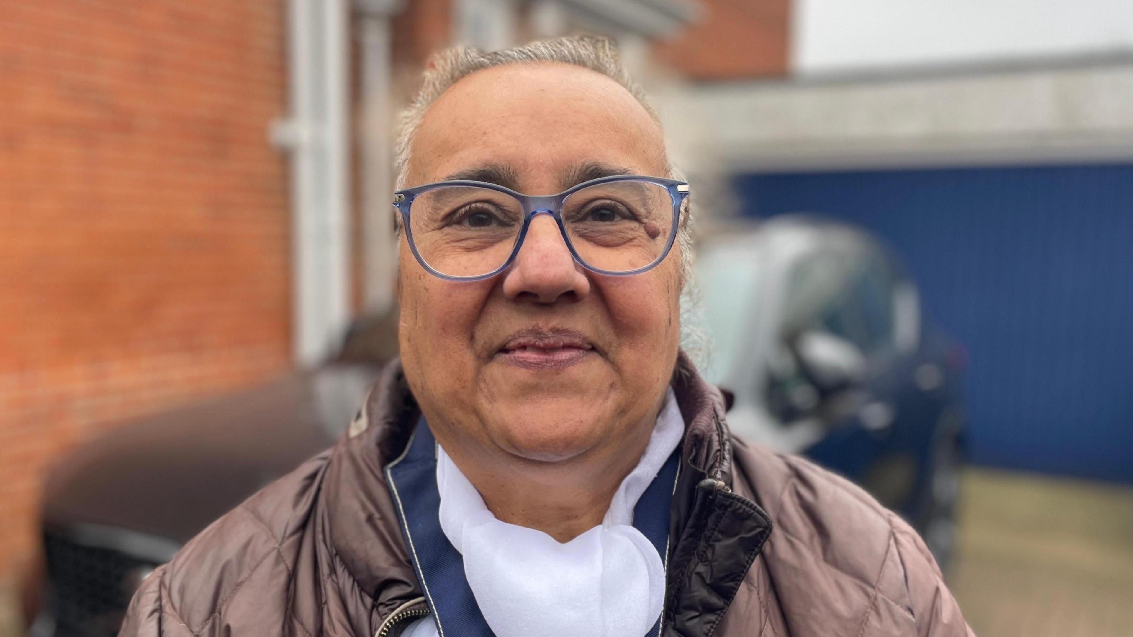A woman smiles at the camera in a head and shoulders image. She is wearing blue-framed glasses and has grey hair that is scraped back in a bun. Her unzipped jacket is quilted and a greyish pink, with a white and blue top underneath.
