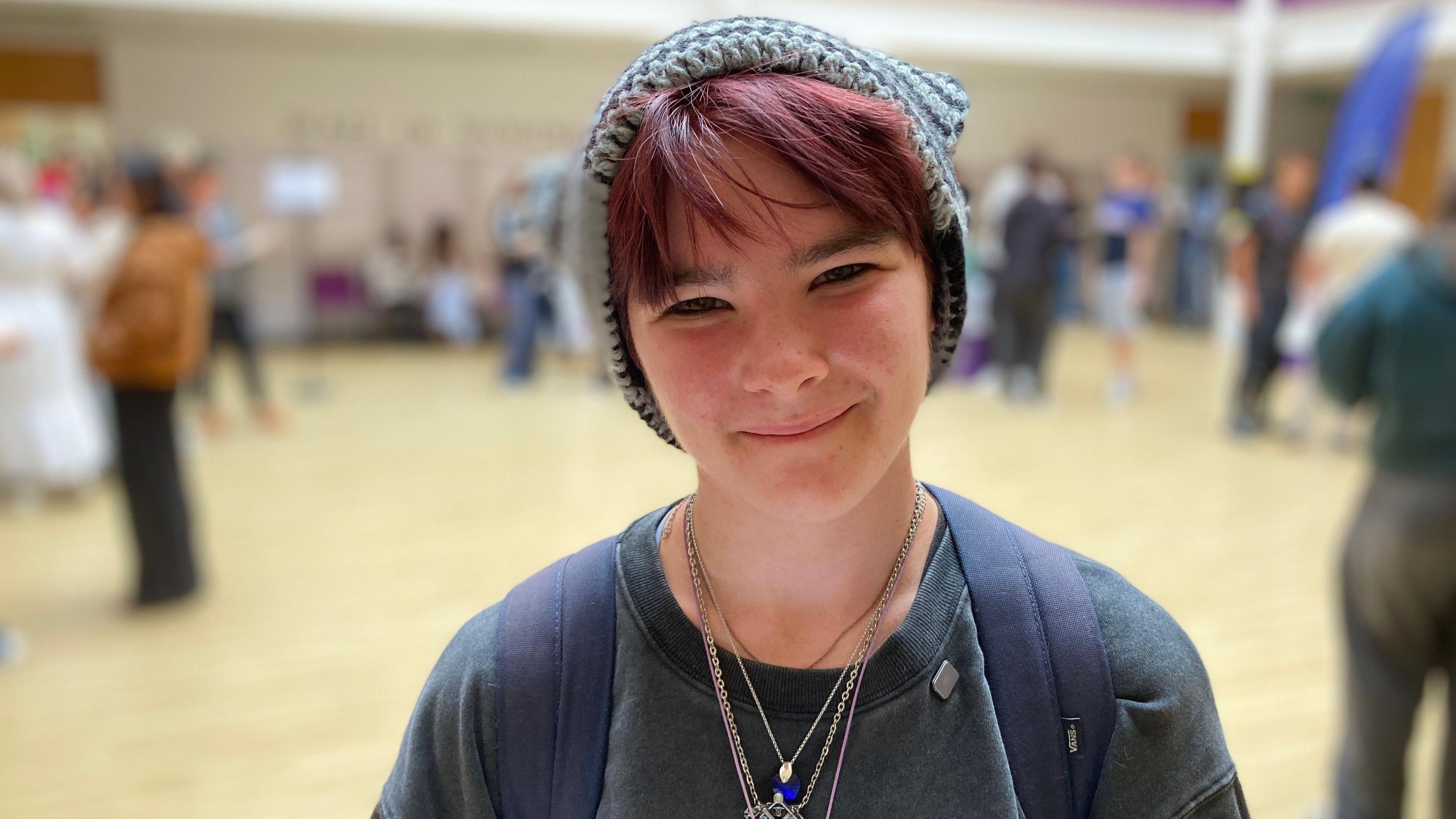 Freya smiles at the camera. She is standing in the school hall, which is blurred behind her. She has red died hair with a long sweeping fringe and wears a grey knit beanie with a dark green sweatshirt and a stack of silver chain necklaces.