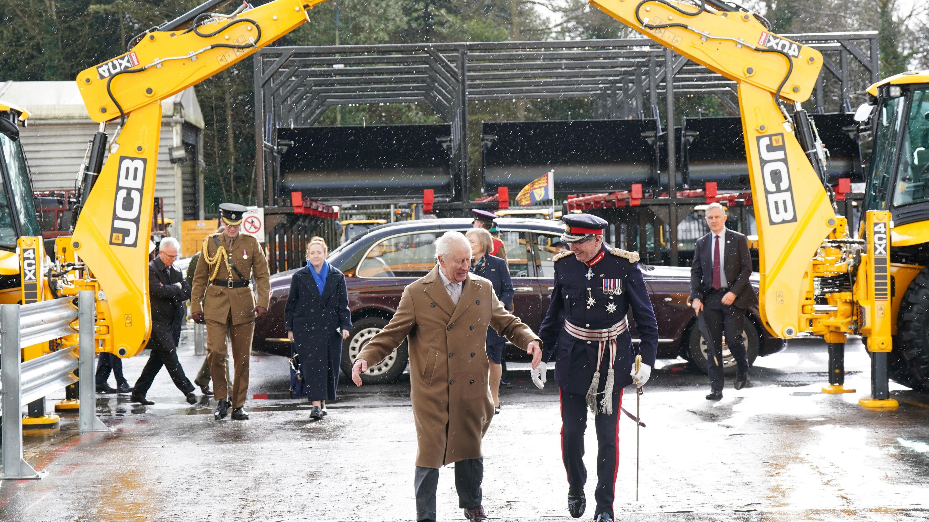 King Charles III, wearing a long brown coat, and is contingent walk underneath an arch created by the arms of two JCB vehicles. A dark red saloon car can be seen in the background.