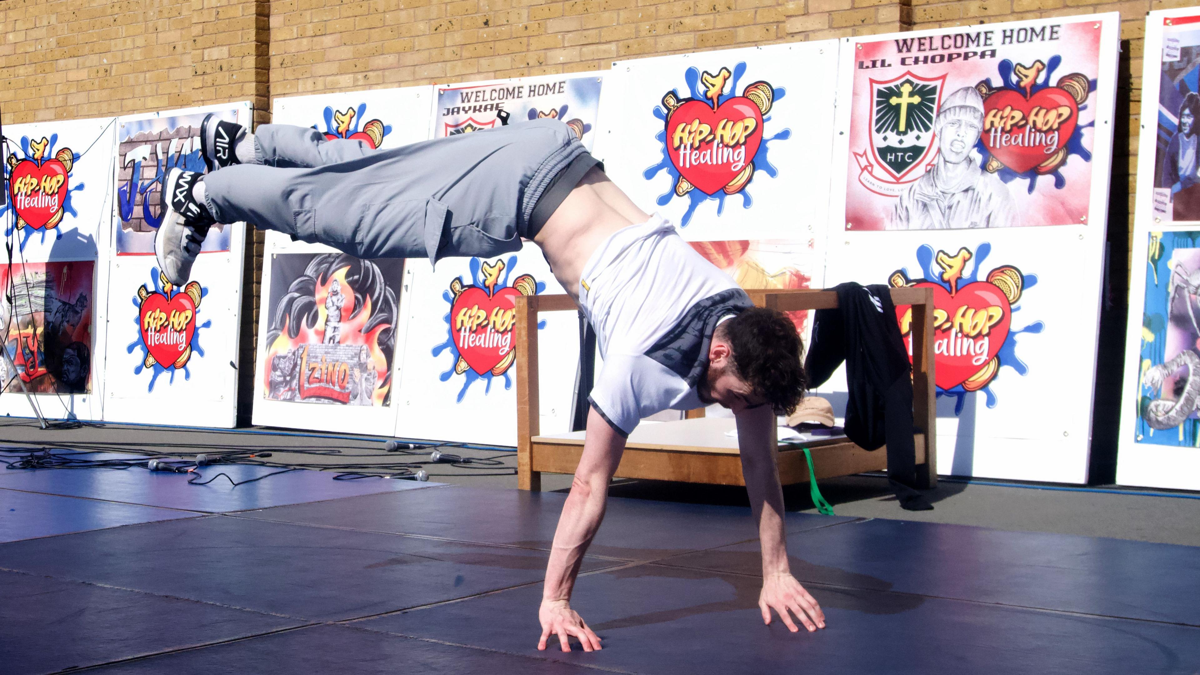 A breakdancer wearing grey trousers and a grey/white t-shirt balancing on his hands with his legs outstretched behind him.