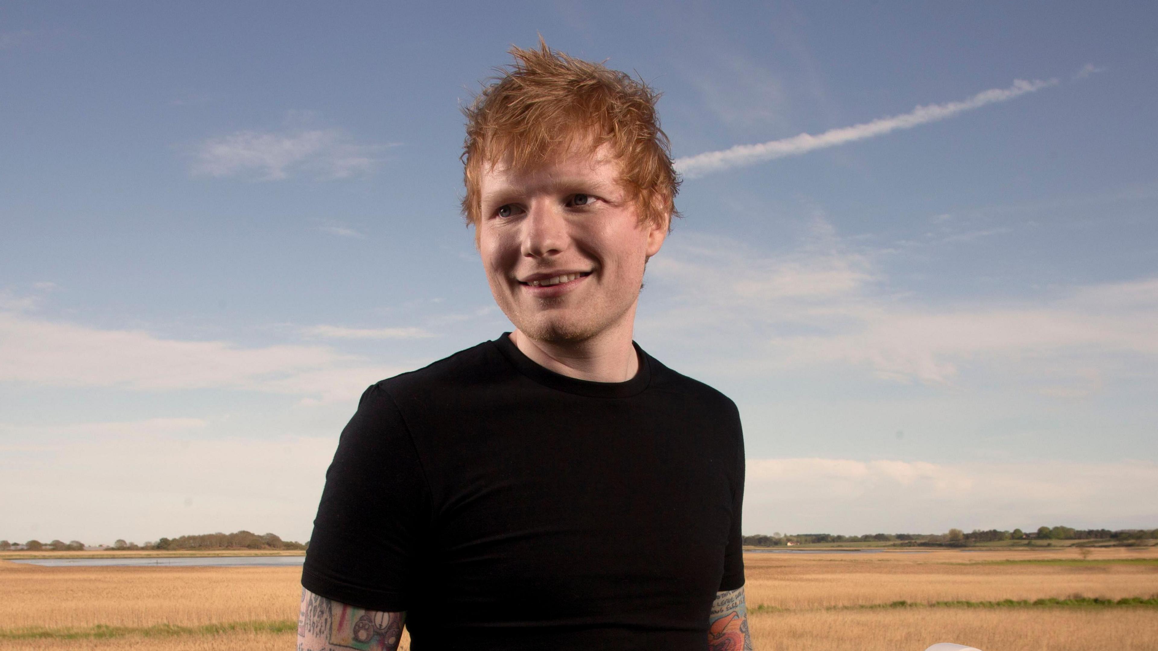 Ed Sheeran standing in front of a white fence in a field in Snape. The ginger haired singer is wearing a tight black T-shirt. His arms are covered in tattoos.