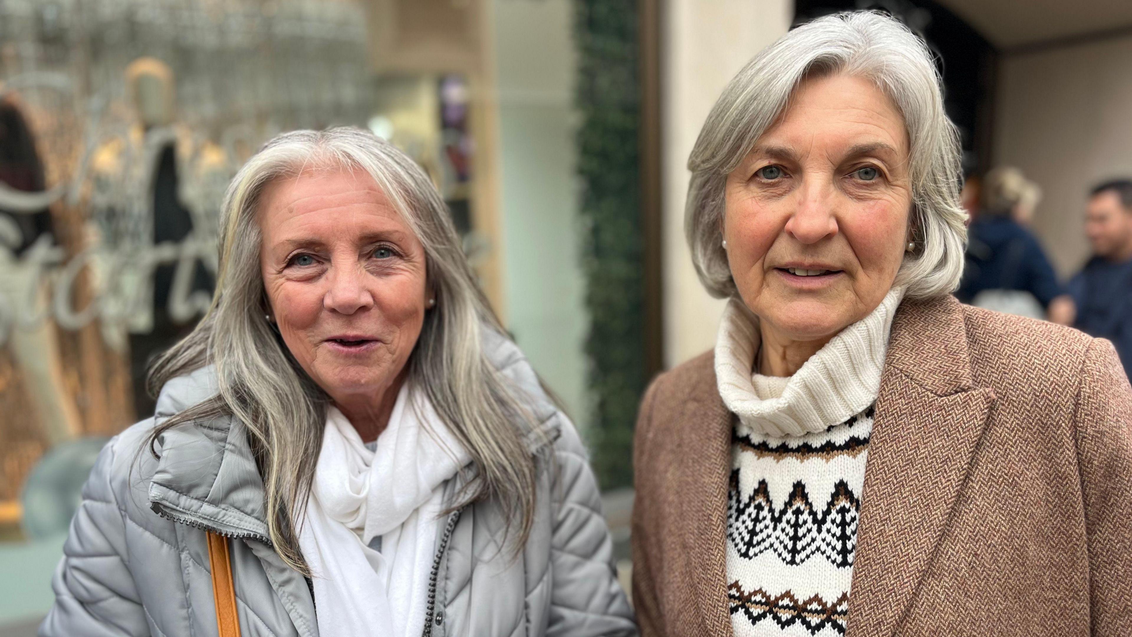 Louise Jackson and Jill Charlton are women with grey hair stood next to each other on a street. Louise is wearing a grey puffer coat and white scarf. Jill is wearing a white turtleneck and a brown tweet coat. 