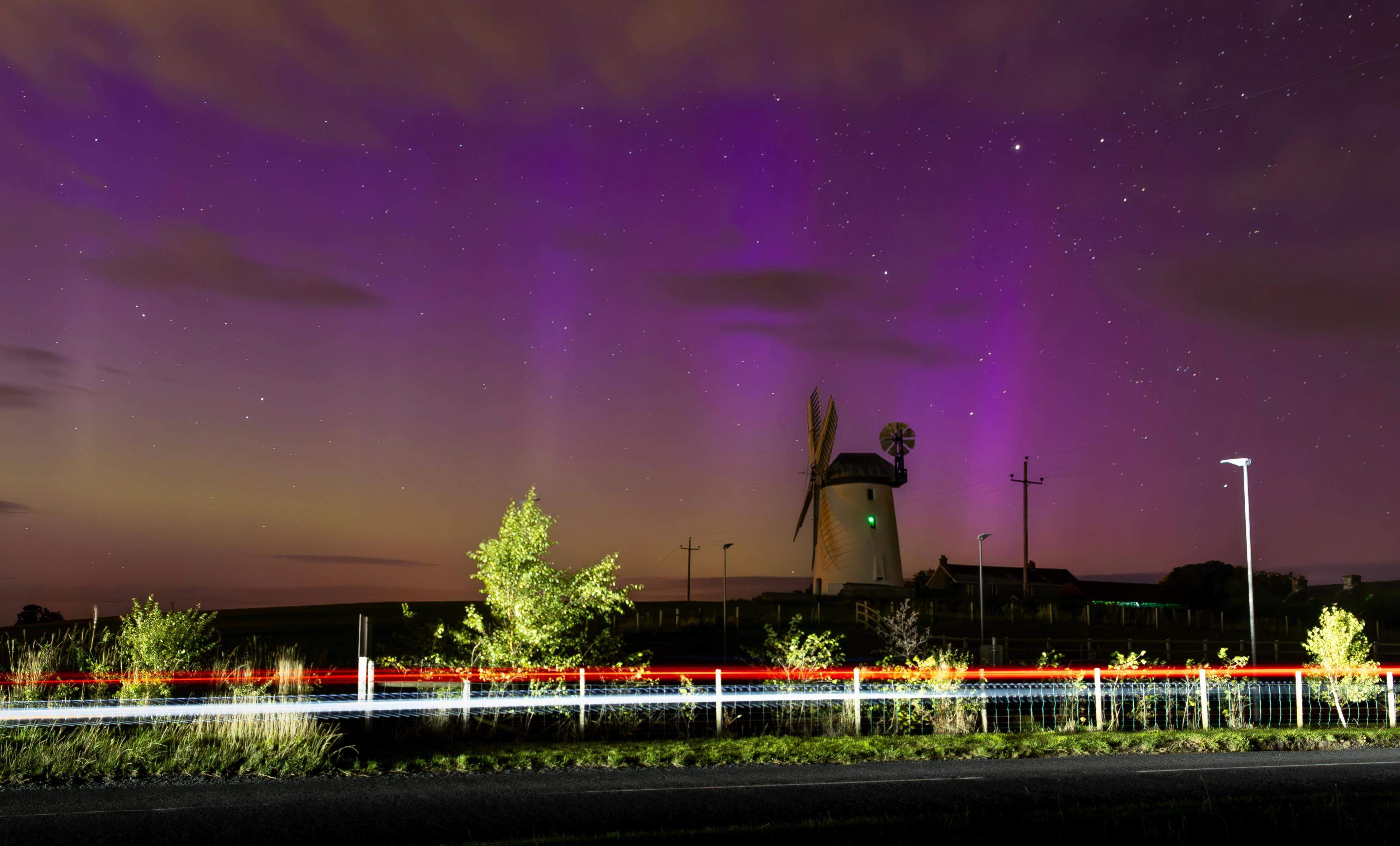 In the foreground some lights from a car passing illuminate some trees at the side of the road. In the background a windmill with purple lights in the sky over it 