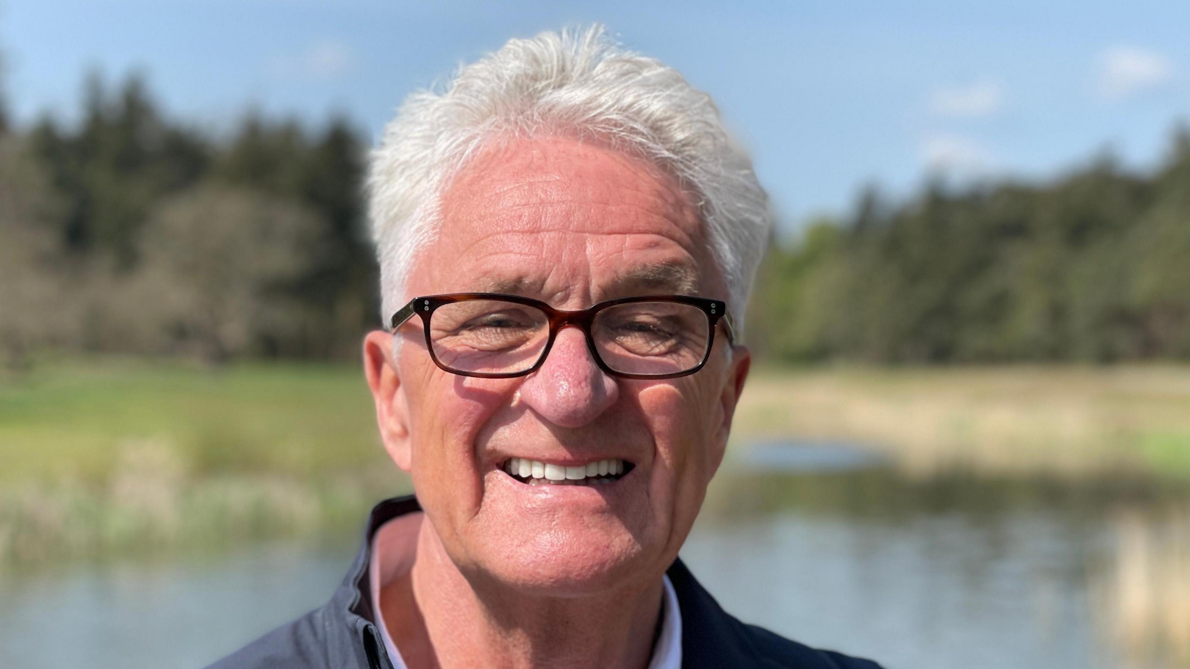 Melvin Benn smiles at the camera on the site of the Latitude Festival. He has white hair and wears glasses and a dark jacket. It is just his face in the shot. Behind him, there is a lake and some trees. The background is out of focus.
