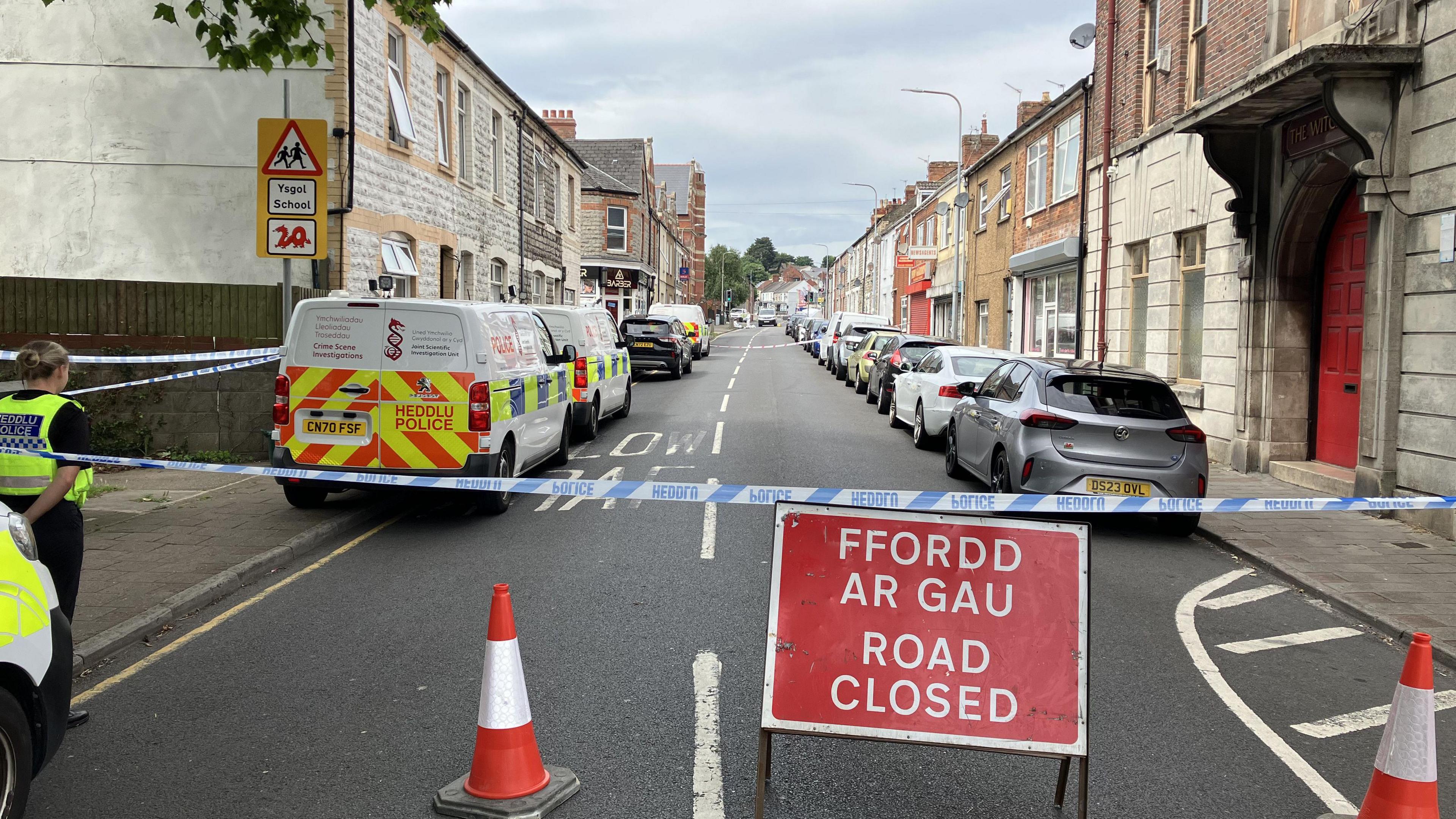 A street blocked off by police tape and cones. You can see this is the case at either end of the road. There is a sign which says FFORDD AR GAU/ Road CLOSED. Police vans line the left side and an officer is visible.