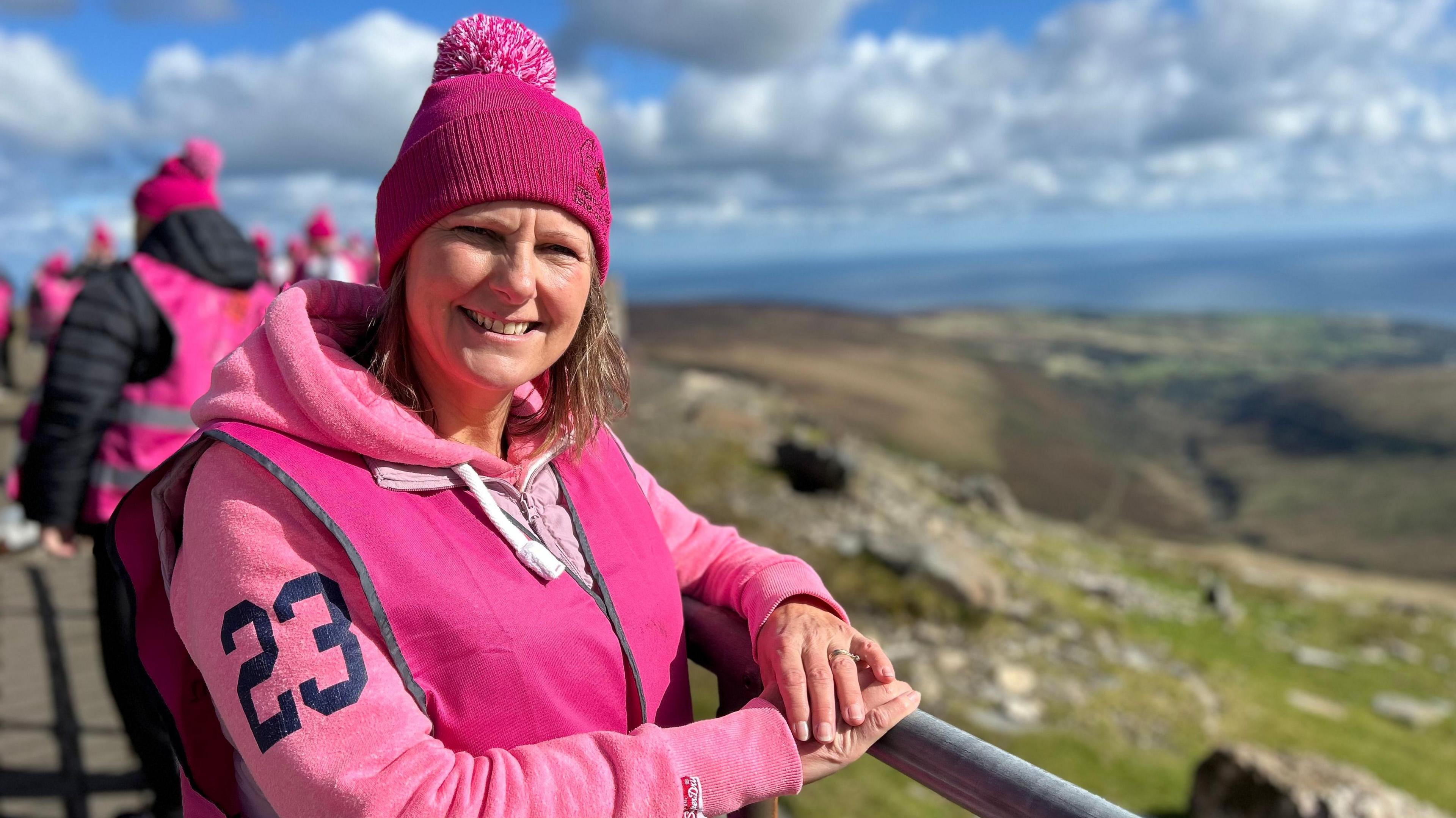 Diane Taylor smiling in front of the rolling Manx hills on a sunny day wearing a pink jumper and bobble hat.