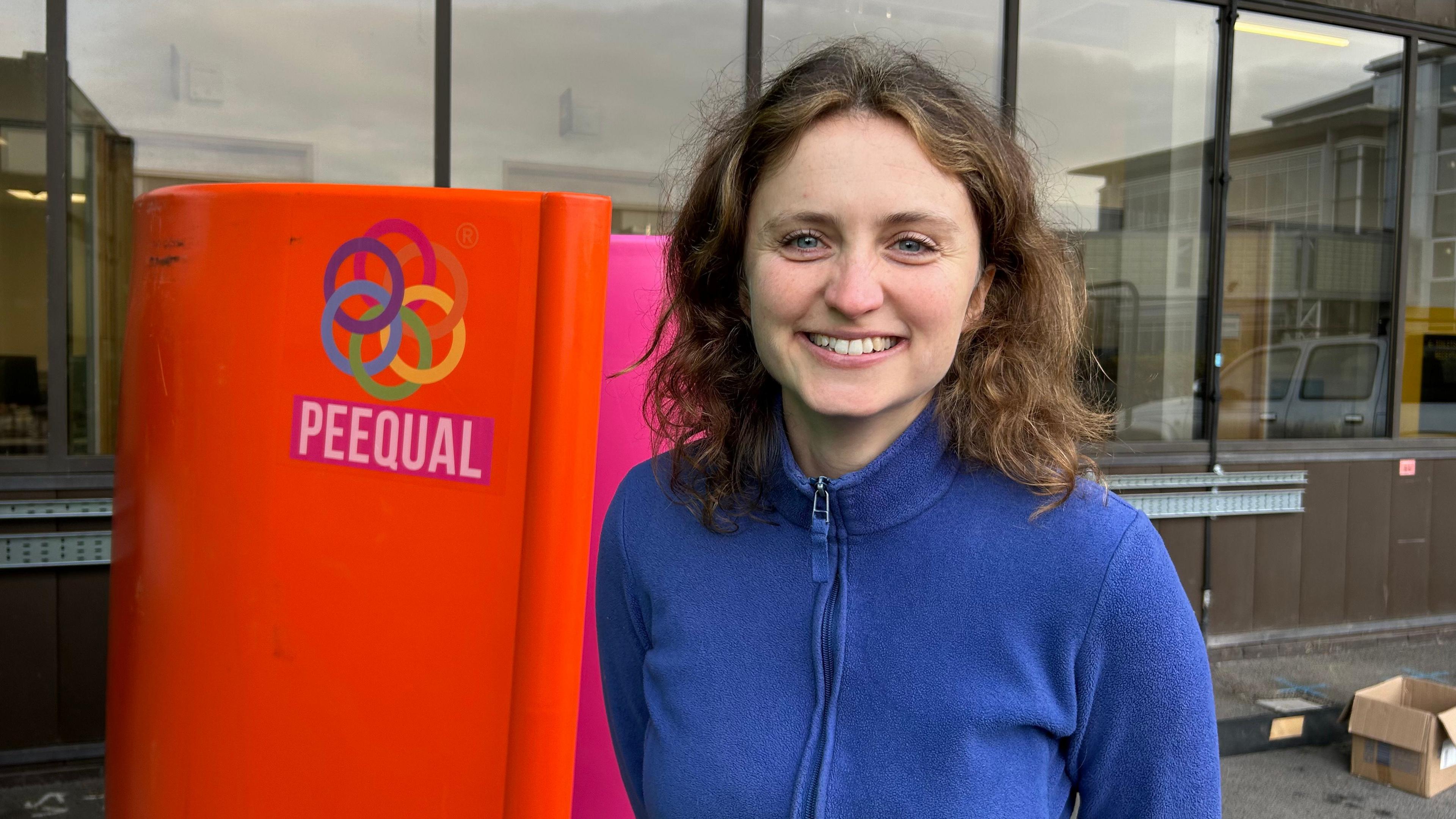 Hazel McShane standing in front of a segment of a PEEQUAL urinal. She has shoulder-length brown hair and is wearing a blue zip-up fleece