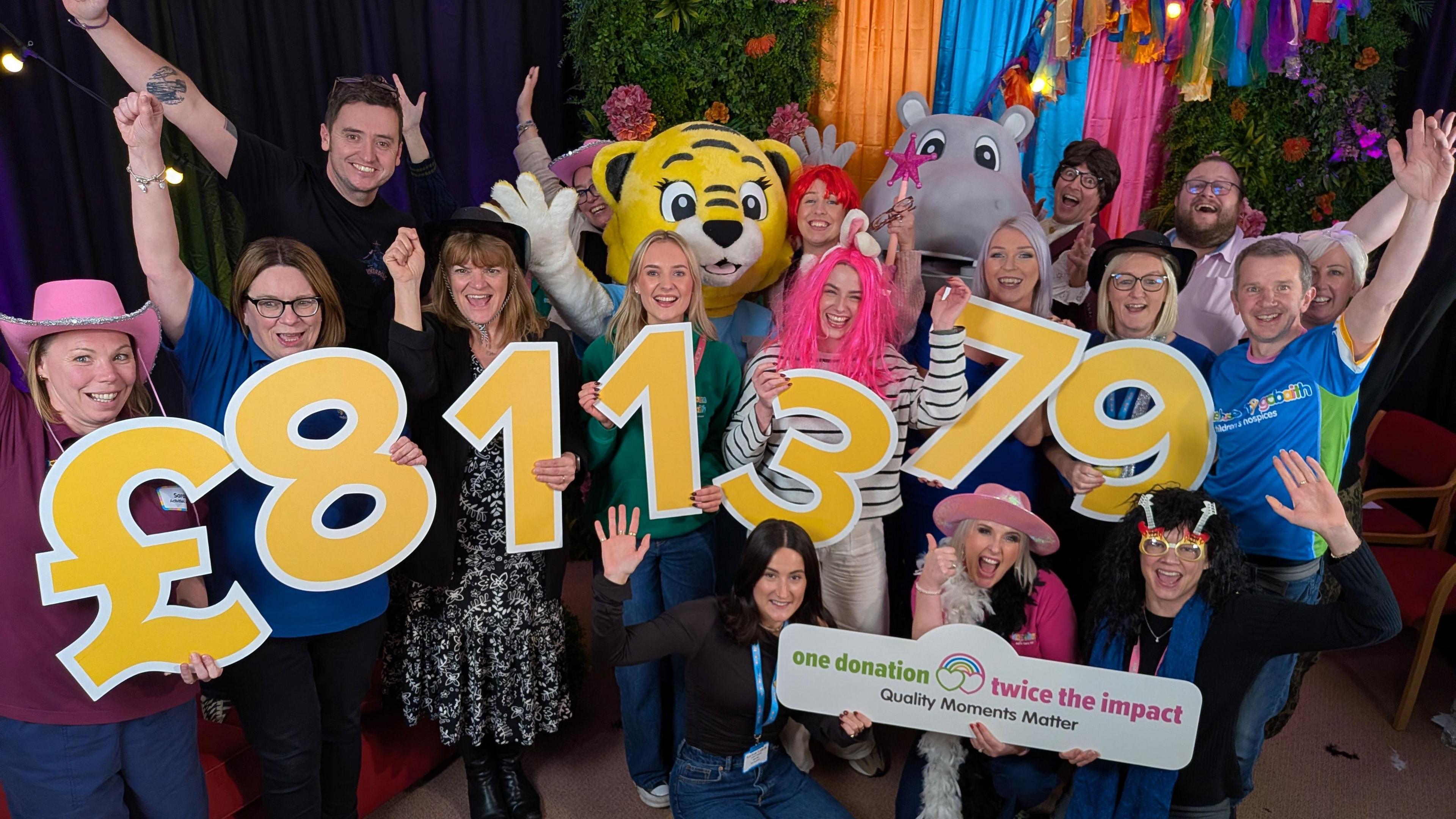 A group of people are celebrating and smiling with their arms raised in the air. Among them is a large tiger costume and a large hippo costume. Between them, they are holding up large yellow numbers that create the number £811,379
