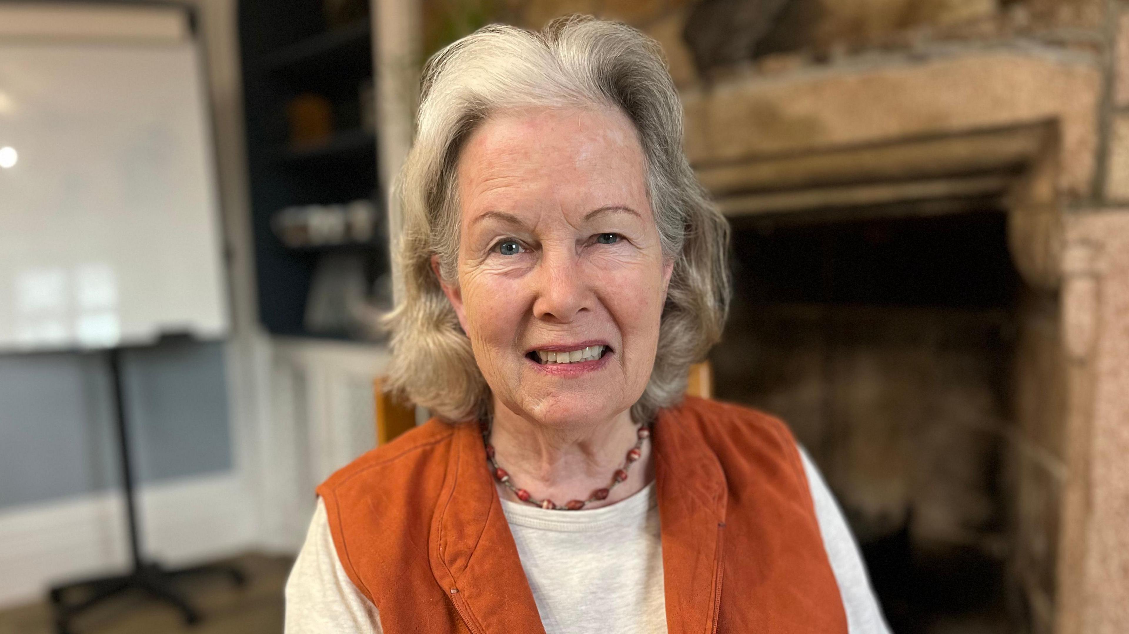 Lee Durrell is looking into the camera, smiling. She is in a room with a fireplace and a flip-chart behind her.