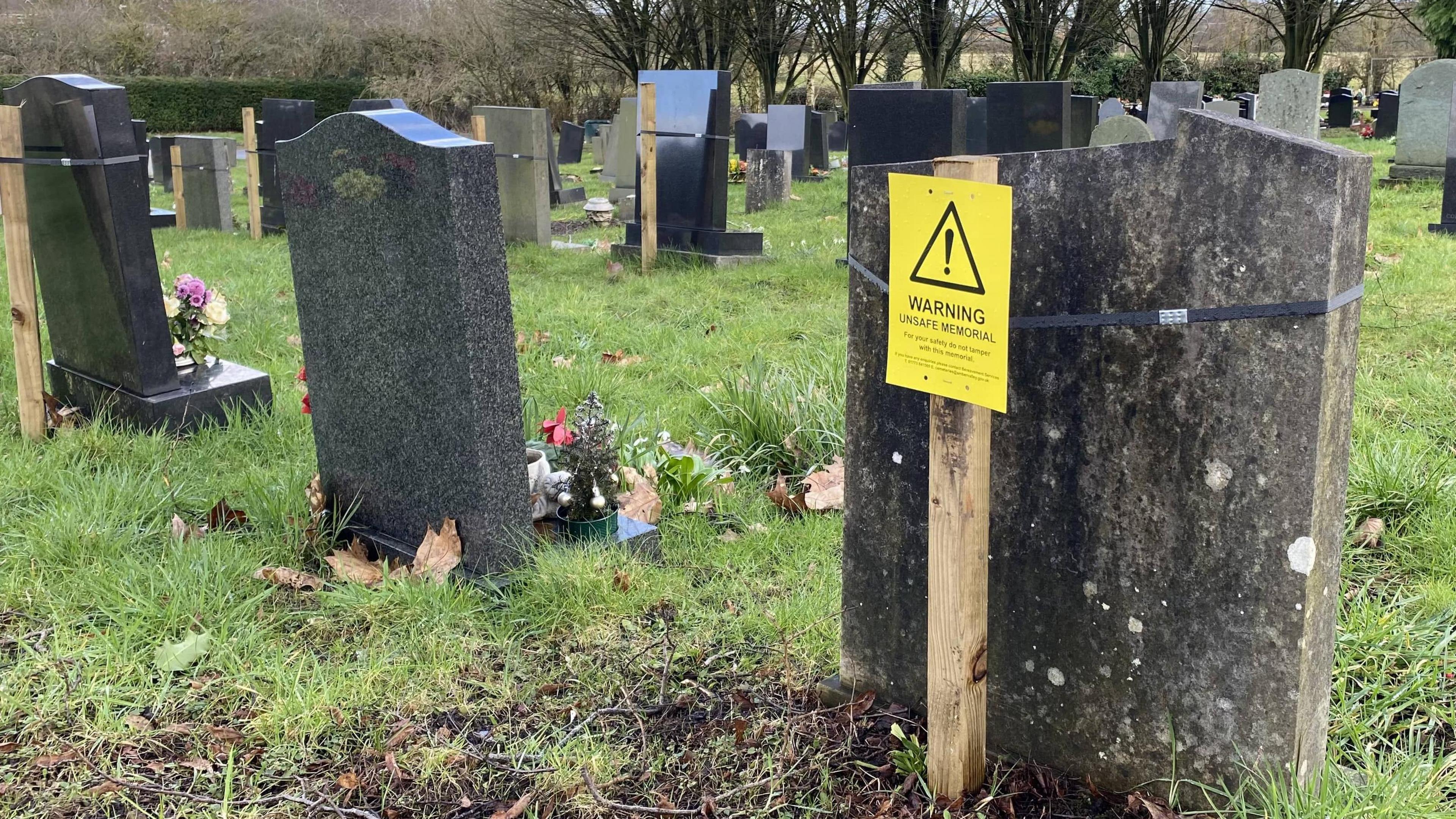 A headstone, with a wooden stick behind it, displaying a yellow 'Warning, Unsafe Material' label