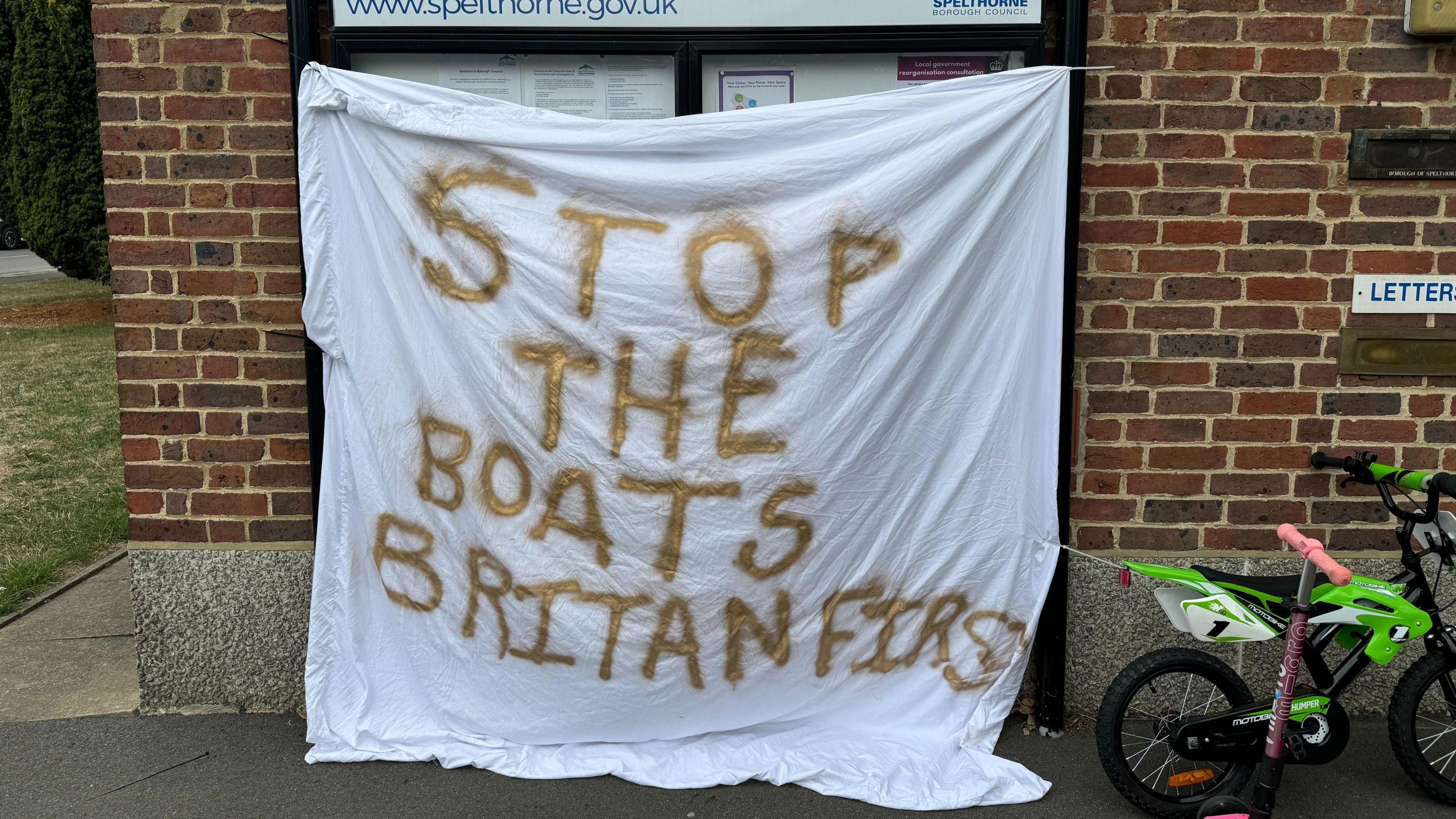 A white bed sheet is tied over a Spelthorne Borough Council noticeboard, with the words "Stop the boats. Britain First" sprayed in gold paint on it. "Britain" is spelt incorrectly as "Britan".