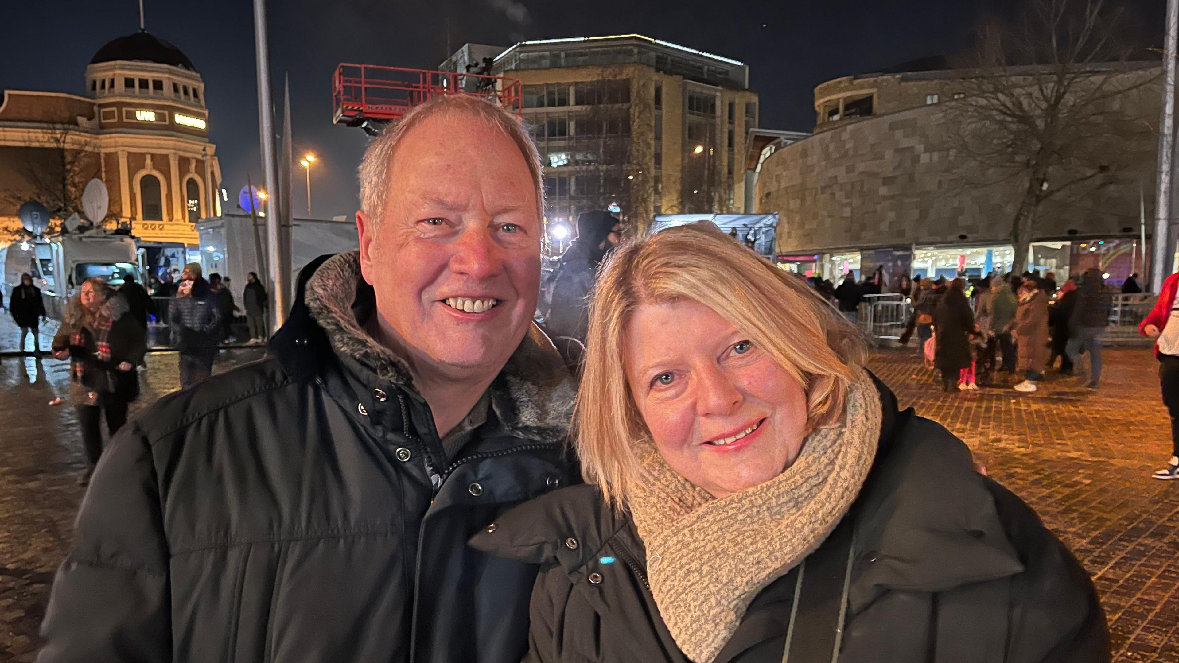 A man and a woman in black puffer jackets standing in Bradford's City Park. She is blonde and wearing a grey scarf. He has short white hair.