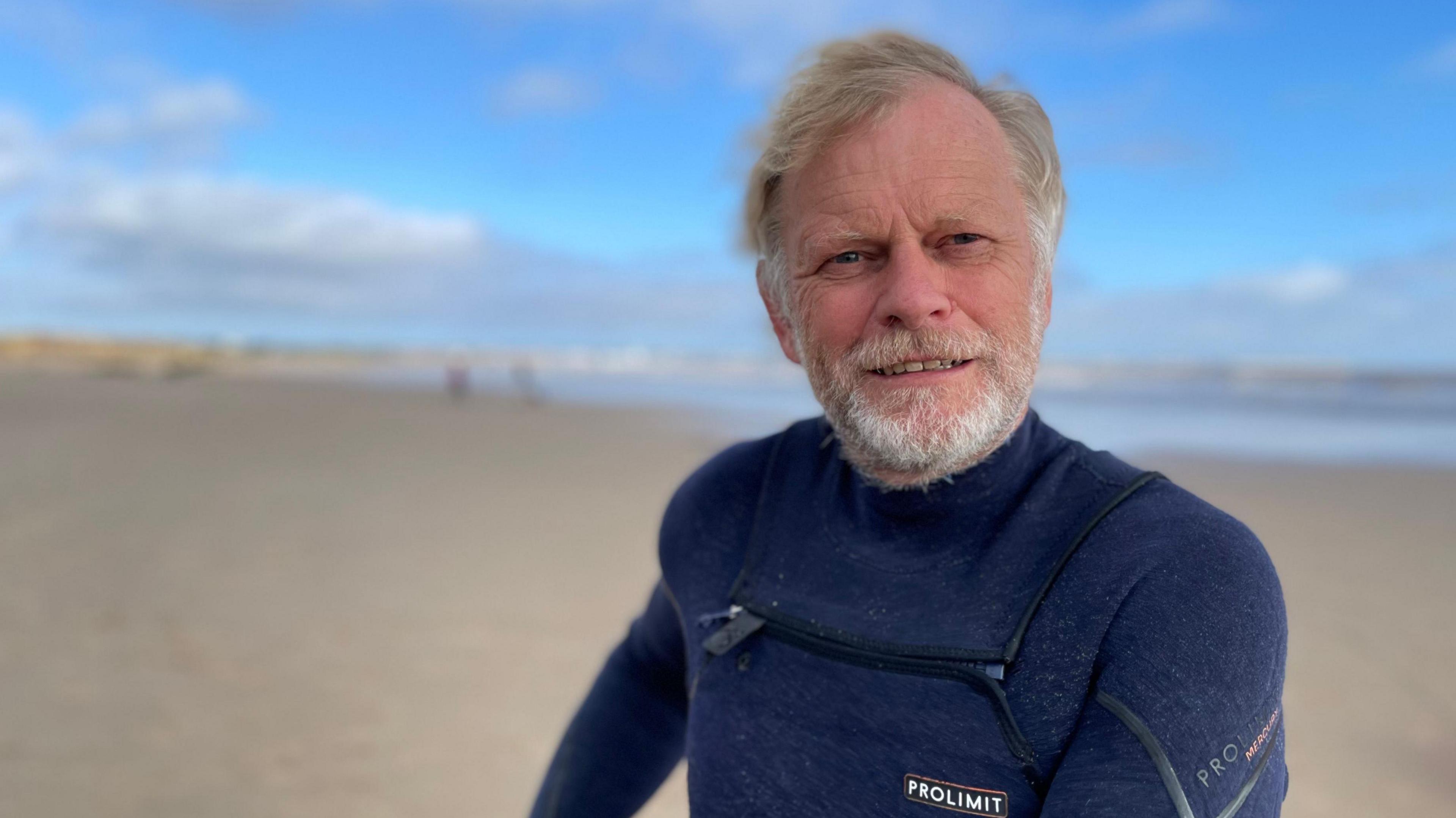 A middle-aged man wearing a blue wetsuit stands on a sandy beach under a blue sky. He has fair hair, which is blowing in the wind, and a white beard 