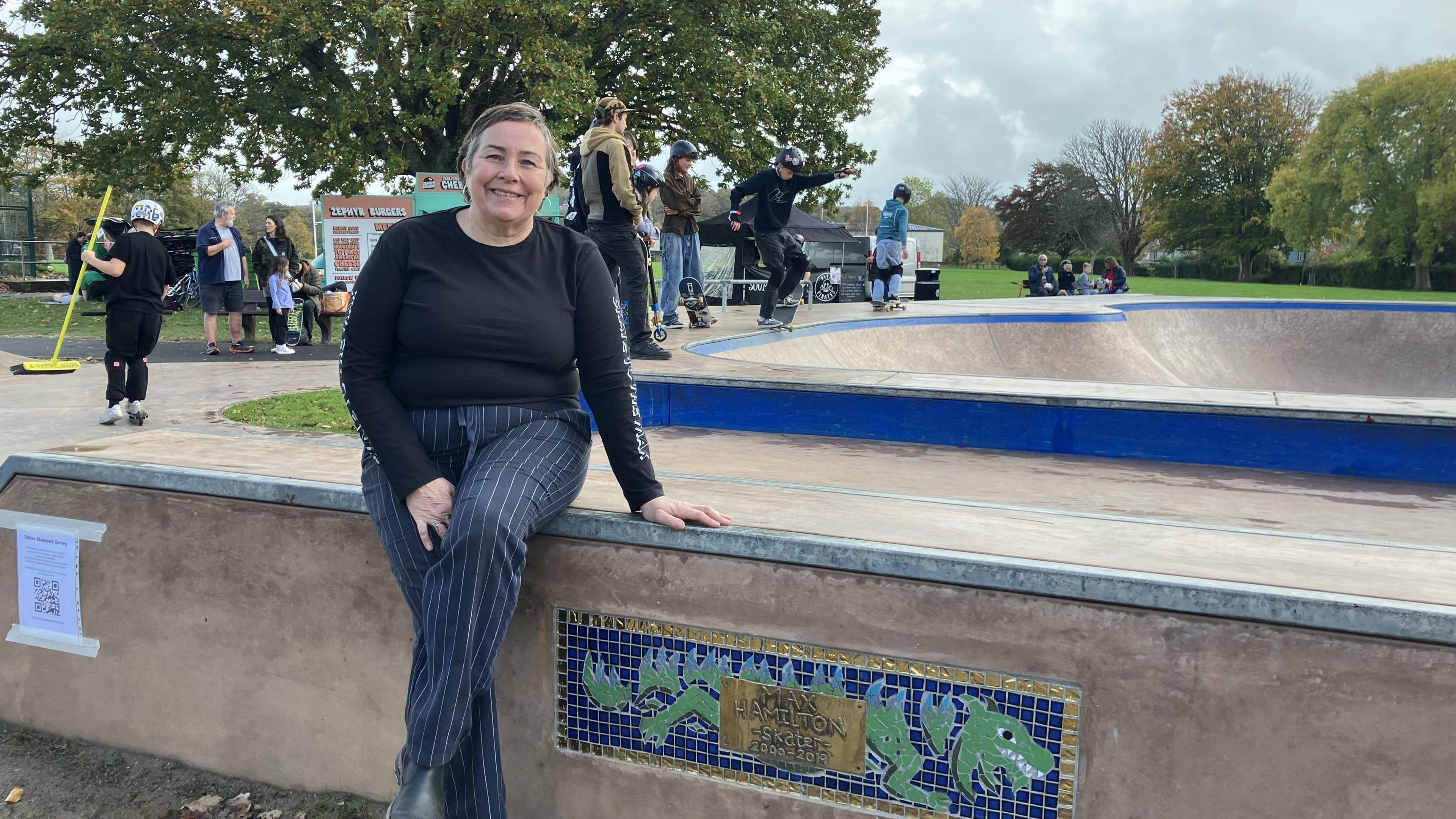 A woman with short brown hair, wearing a long sleeve black t-shirt and pinstripe navy blue trousers. She is sat on a concrete block in a skatepark in front of a concrete skate bowl. There are children wearing helmets riding skateboards and scooters in the background.