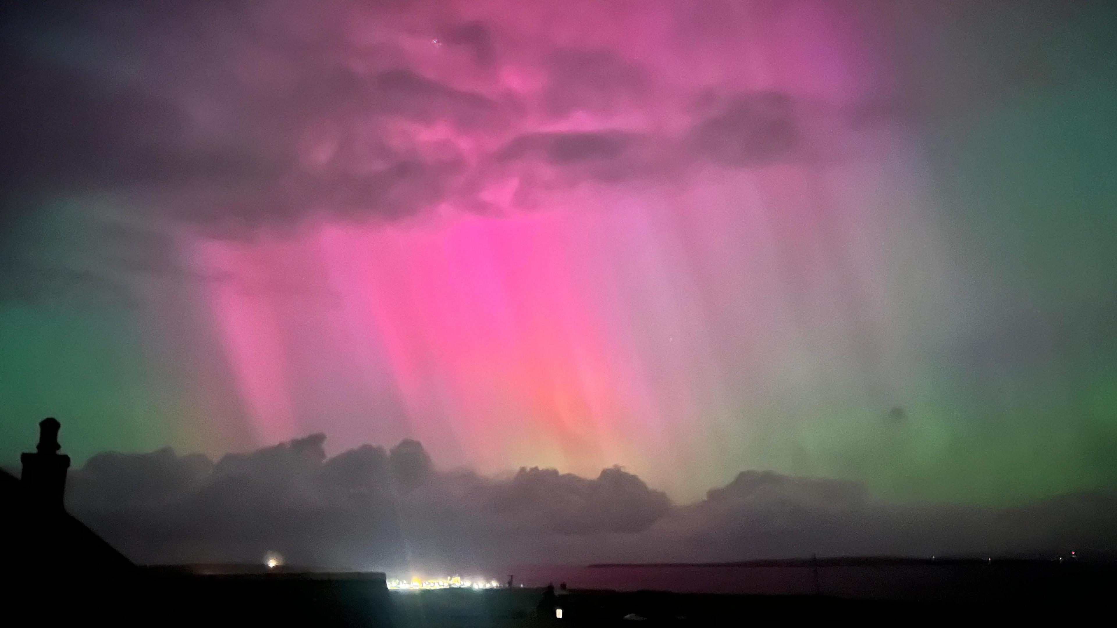 Pink and green aurora shine through clouds amid a dark sky over a rural coastline