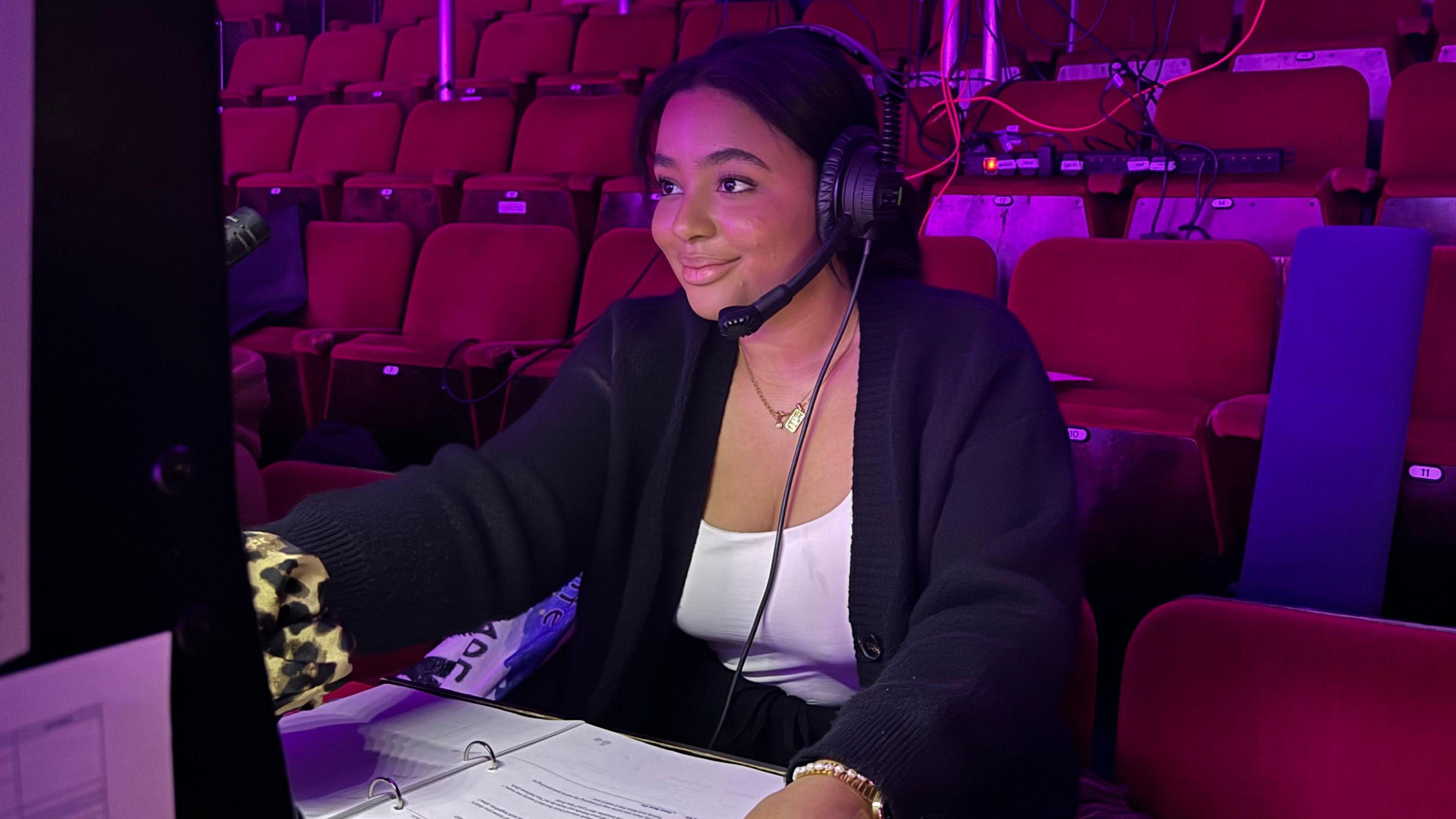 Emily Obasahan sitting at the stage manager's station in the theatre. She has a headset on and is wearing a white top with a dark jacket and a gold necklace. Her hair is tied back, and she is smiling. She has a folder with instructions in front of her. There are rows of empty  burgundy theatre seats behind her. 