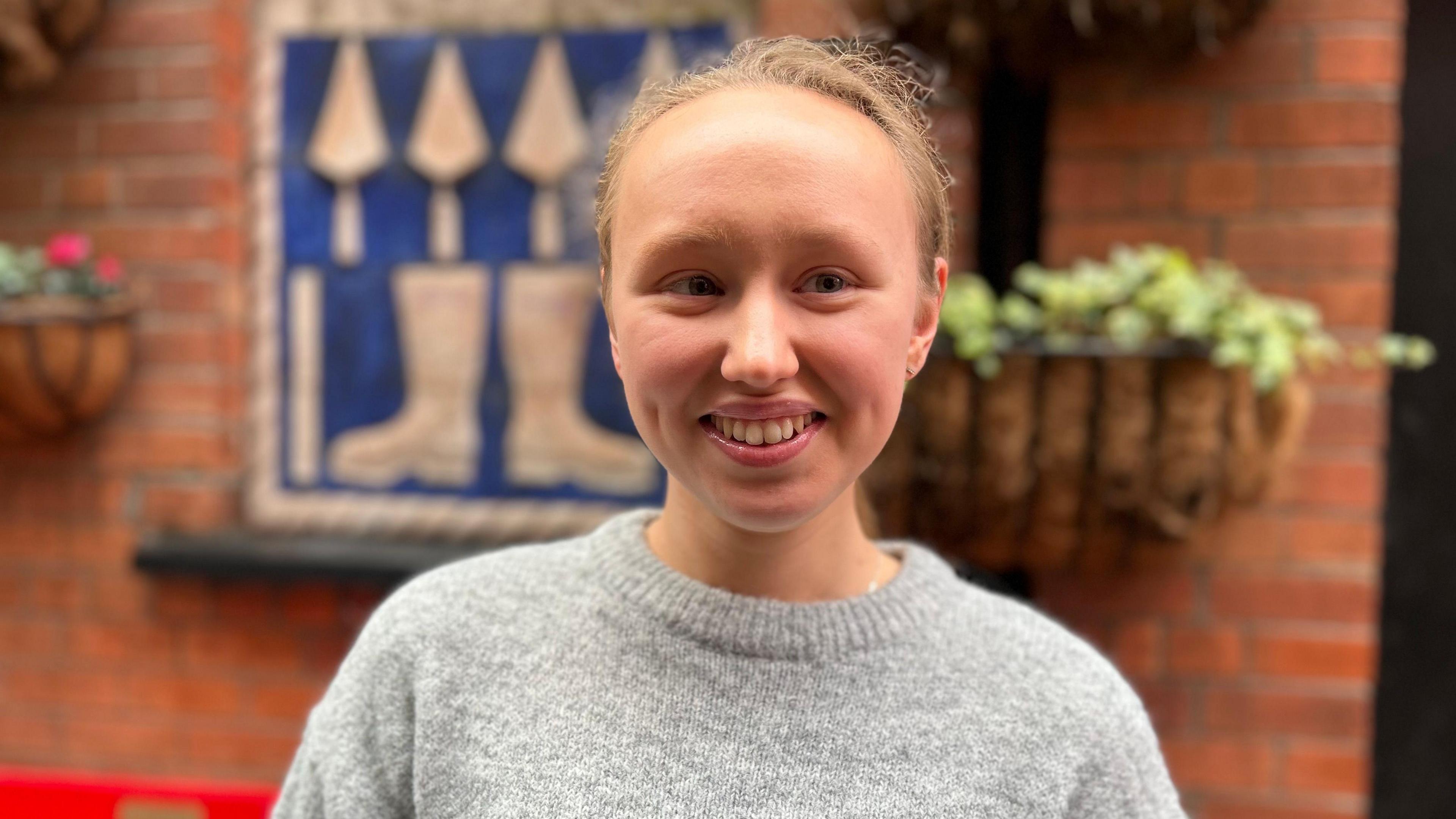 Georgia Hussey in a grey jumper, pictured against a red brick wall, she has blonde curly hair tied up