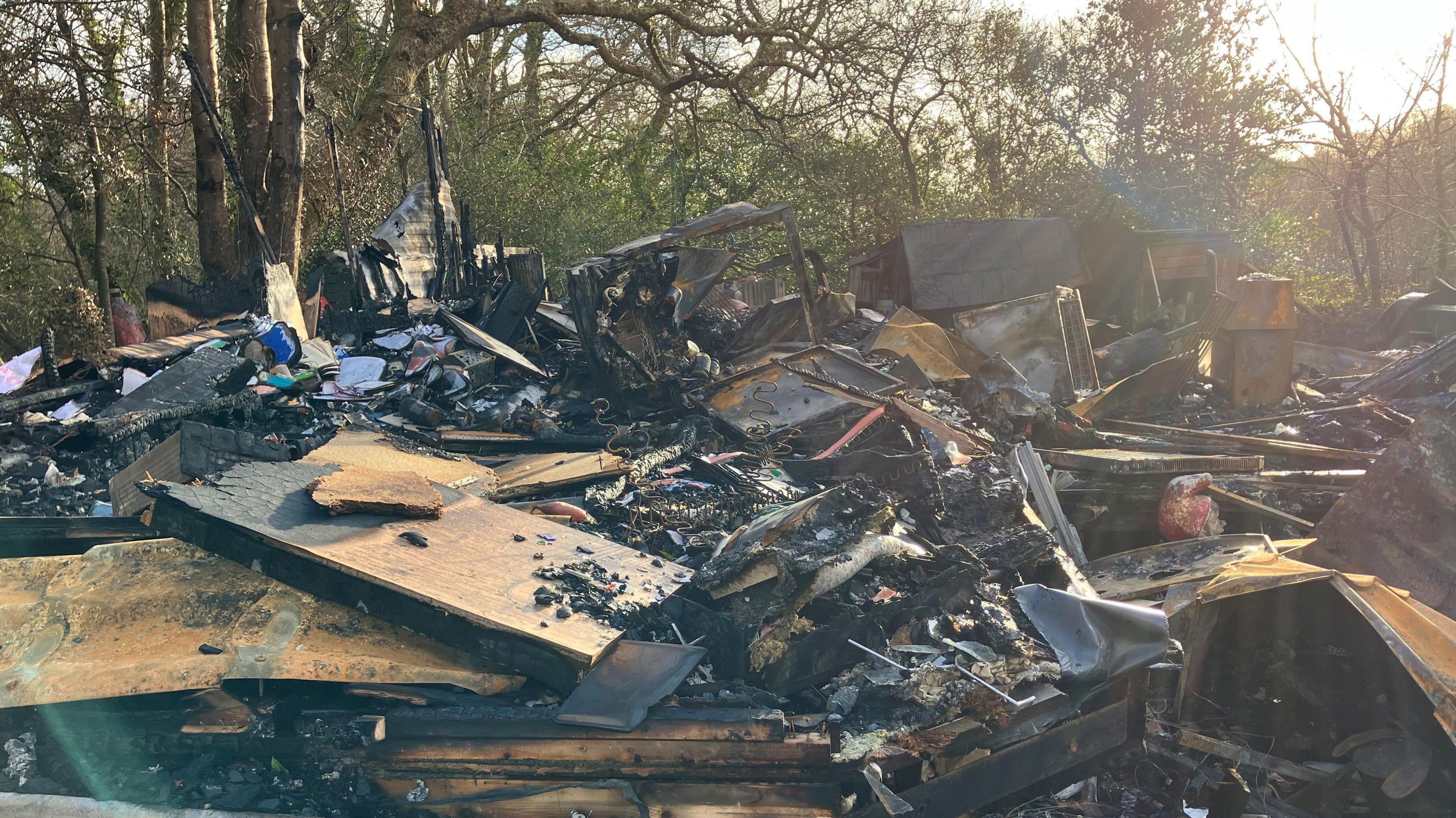 A mass of charred wood and burnt belongings surrounded by trees